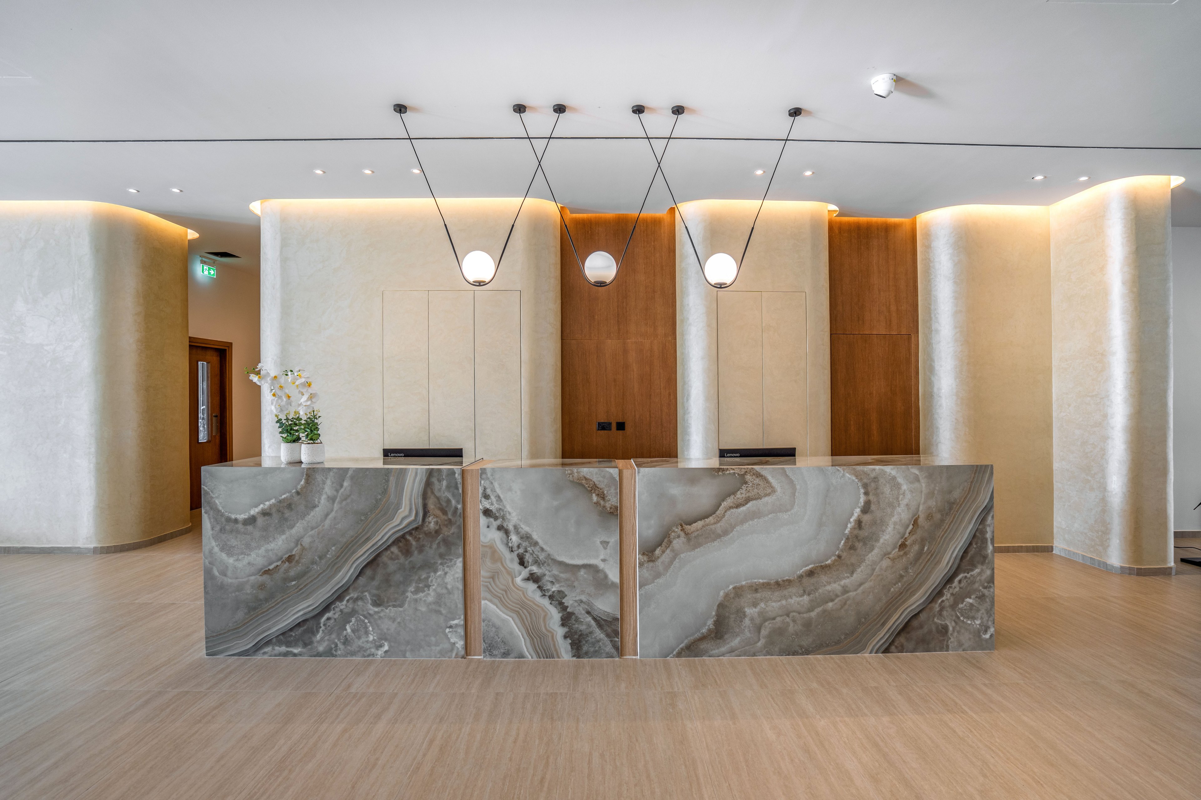 A contemporary reception desk with a striking marble front stands in the center of a minimalist lobby. Soft, indirect lighting highlights the curved cream walls and wooden accent panels behind the desk. Three pendant lights hang above the counter, adding a refined decorative touch. The space appears clean, bright, and welcoming, suggesting a modern hotel or office entrance.