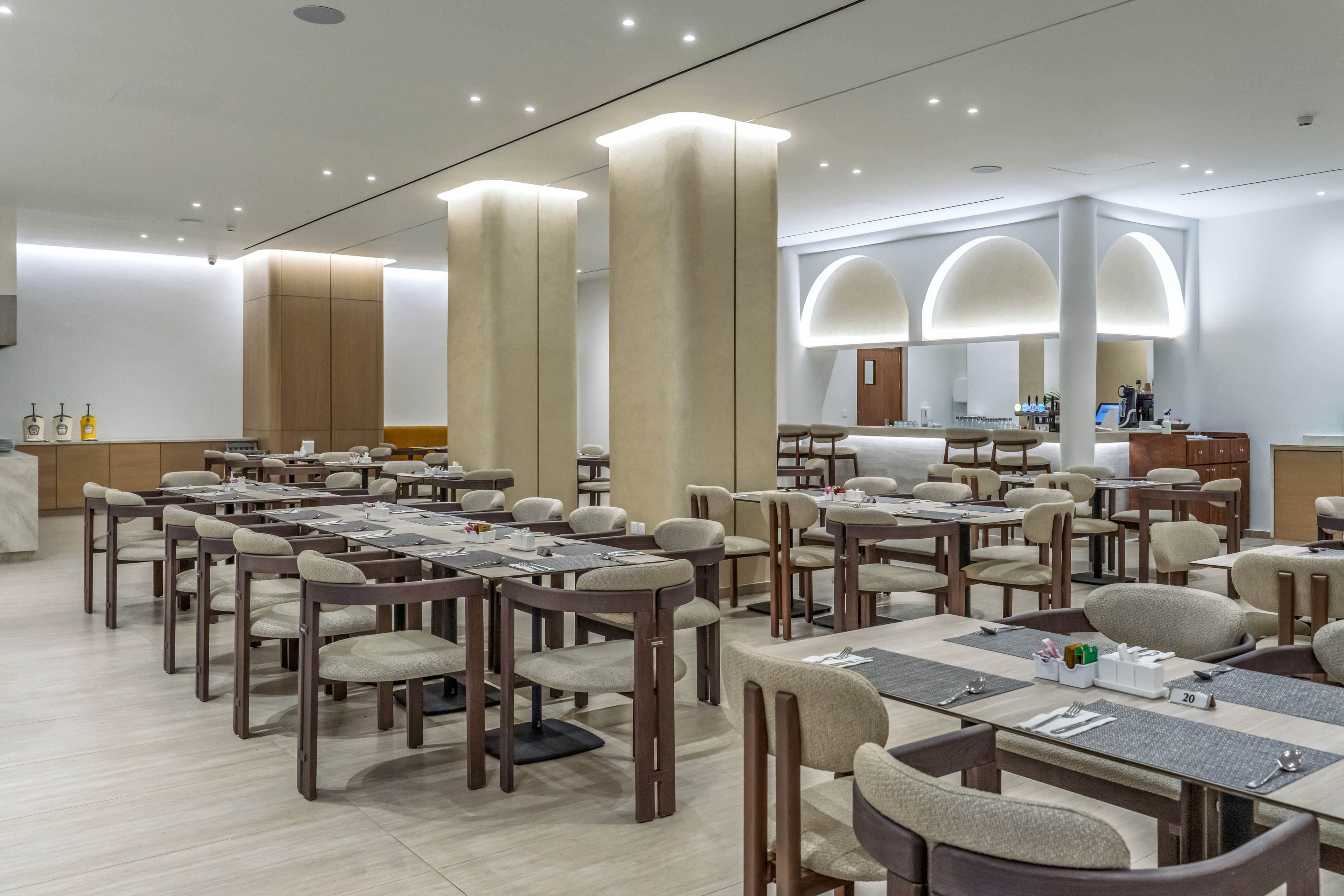 A contemporary restaurant dining room features rows of wooden tables and cushioned chairs arranged neatly. The space is bright and open, with tall columns and recessed ceiling lighting creating a clean, modern look. Neutral tones dominate the palette, including beige floors, light upholstery, and soft white walls. In the background, an illuminated bar or service counter and arched wall niches add architectural interest to the calm, sophisticated setting.