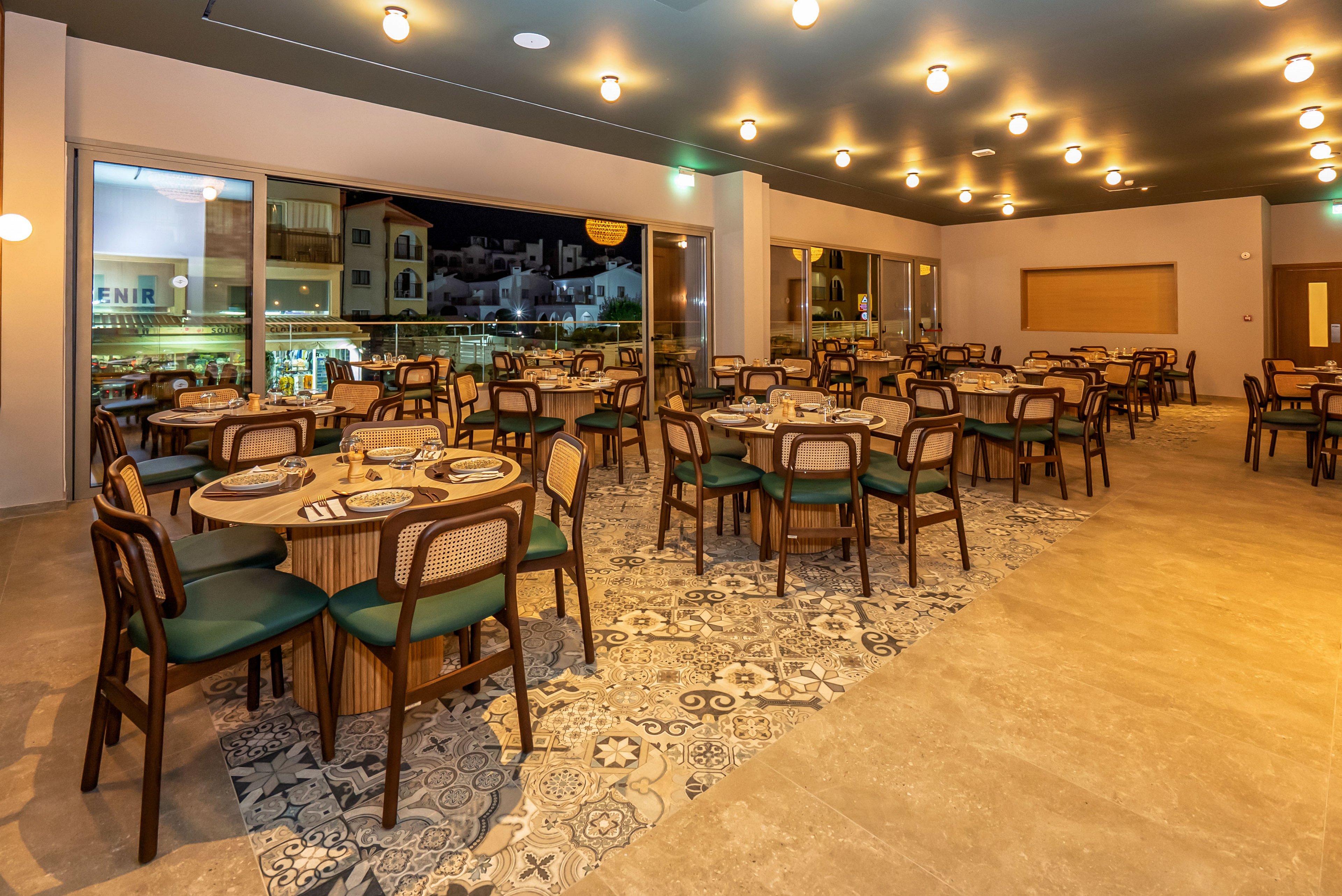 A contemporary restaurant dining area is shown empty at night, with round wooden tables and cushioned chairs neatly arranged. Warm ceiling lights illuminate the space, highlighting a patterned tile floor section contrasting with smooth flooring. Large windows reveal an exterior nighttime street scene with nearby buildings. The overall atmosphere feels clean, inviting, and ready for guests.