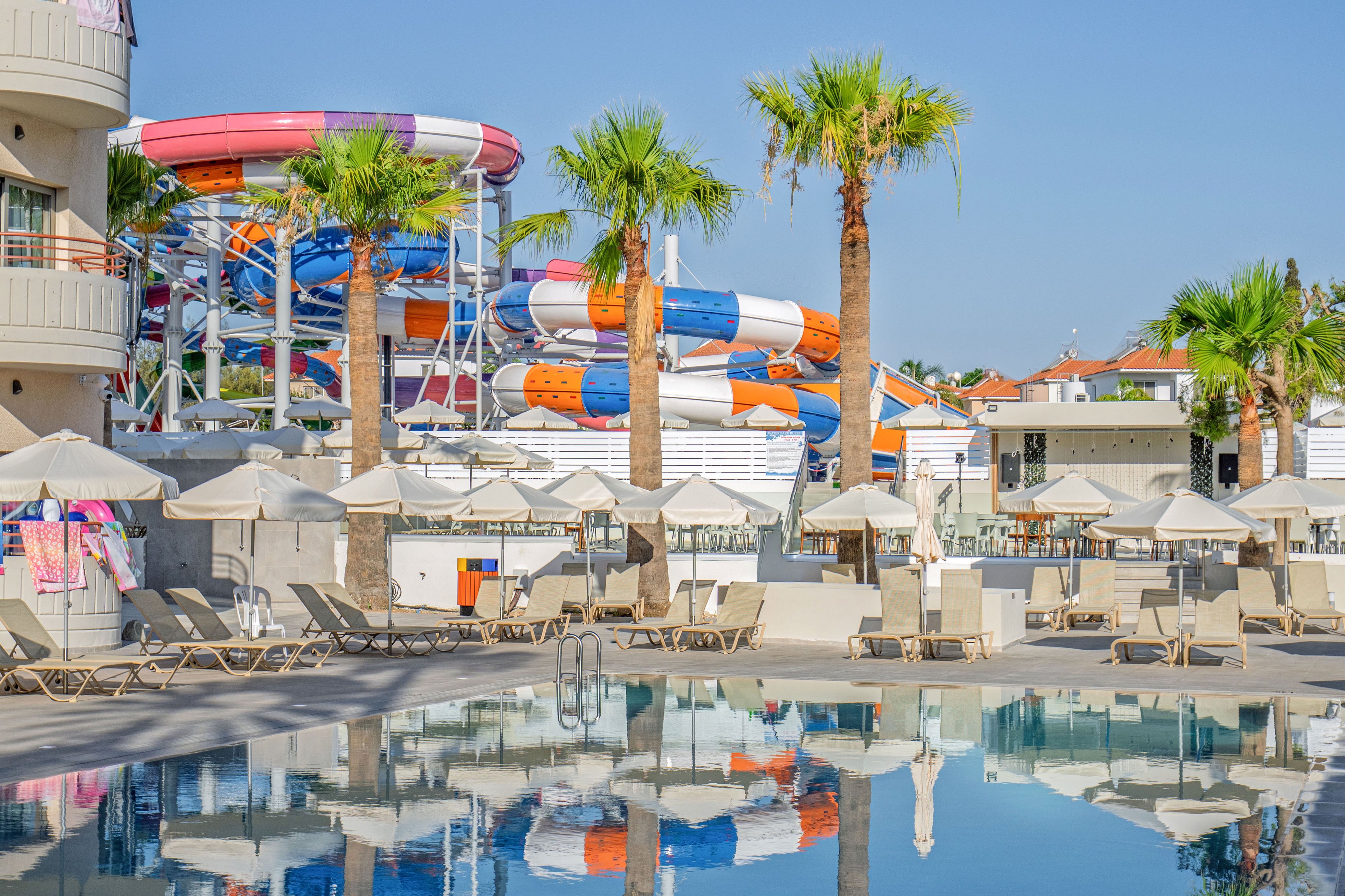 A sunny resort pool area features empty lounge chairs and umbrellas reflected in the calm water. In the background, large colorful water slides in red, orange, blue, and white dominate the scene, suggesting a nearby water park. Palm trees add a tropical atmosphere, and nearby buildings frame the leisure space. The bright daylight and clear sky create a relaxed vacation mood.