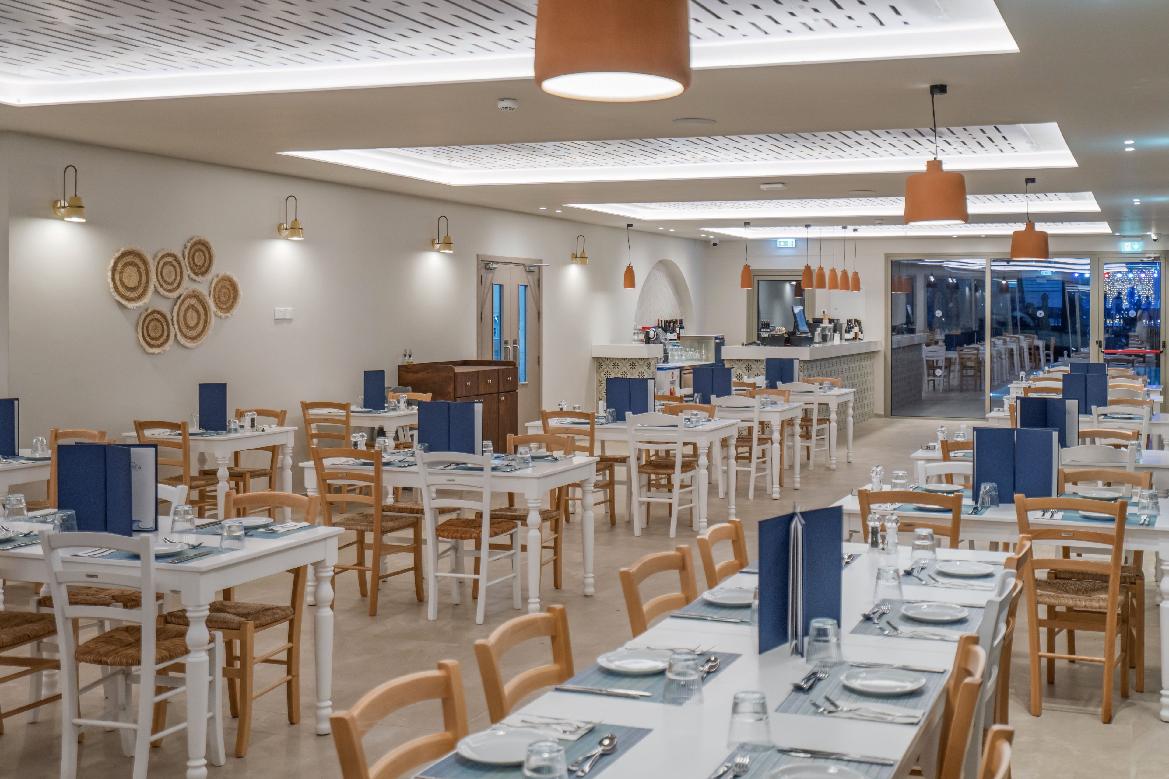 Spacious restaurant dining room with neatly arranged wooden tables and chairs. White and light wood furniture is set with plates, glasses, and cutlery, with blue menu folders adding contrast. Warm pendant lights and wall sconces illuminate the space, highlighting decorative wall art and a bar area in the background. Large windows at the far end suggest an open, airy atmosphere suitable for group dining and events.