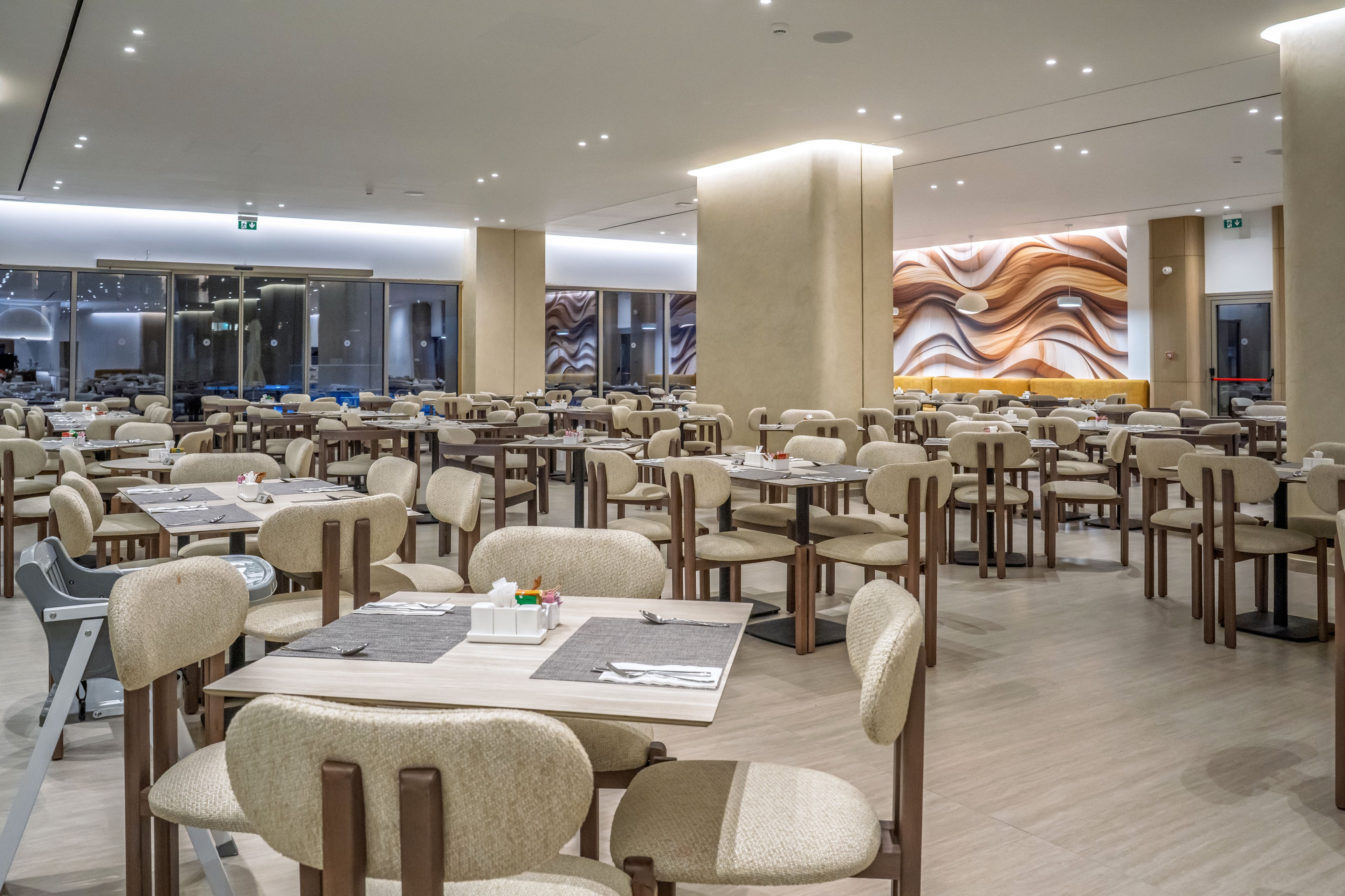 Spacious contemporary restaurant dining room with rows of tables and upholstered chairs. The setting appears clean and orderly, with neatly arranged place mats and condiments on each table. Warm neutral tones dominate the space, complemented by soft ceiling lighting and large windows along one side. A decorative abstract wall mural adds visual interest and a modern design accent to the interior.
