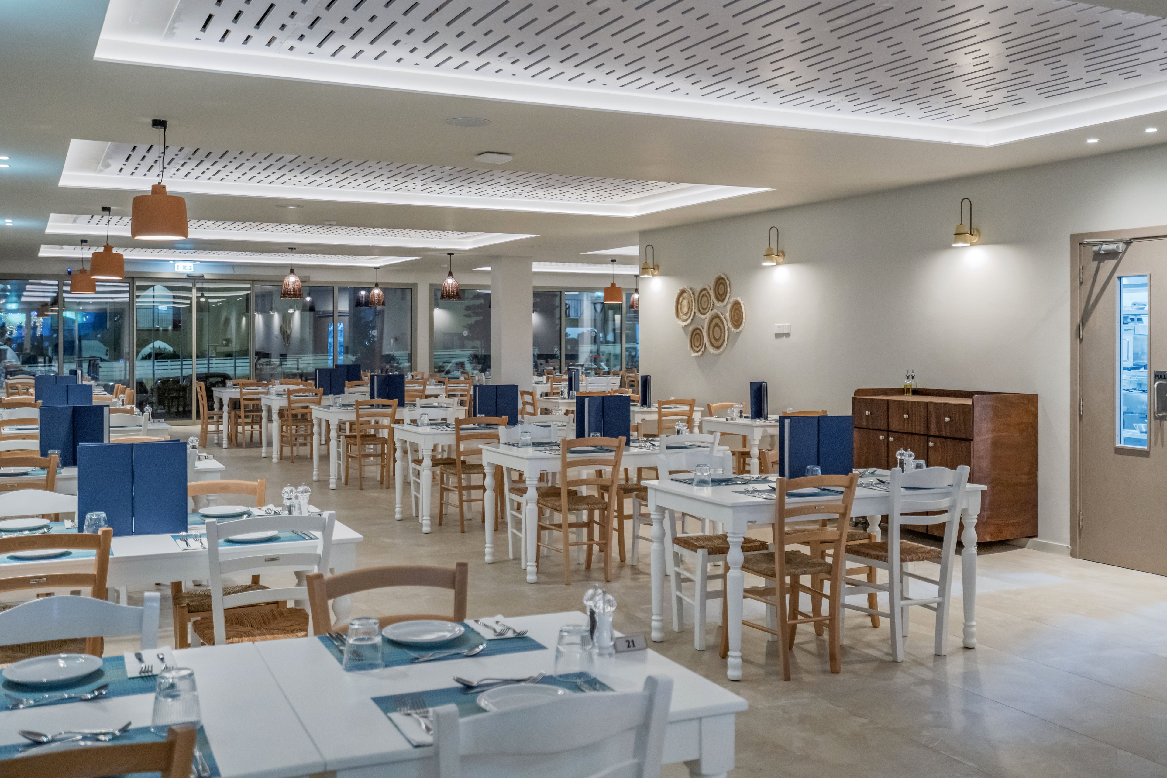 Spacious restaurant dining room with neatly arranged white tables and wooden chairs. Blue table settings with plates and napkins create a fresh, coastal-inspired look. Warm pendant lights and wall sconces illuminate the space, highlighting decorative wall plates and a sideboard. Large windows and glass doors open to an outdoor area, enhancing the airy, contemporary atmosphere.