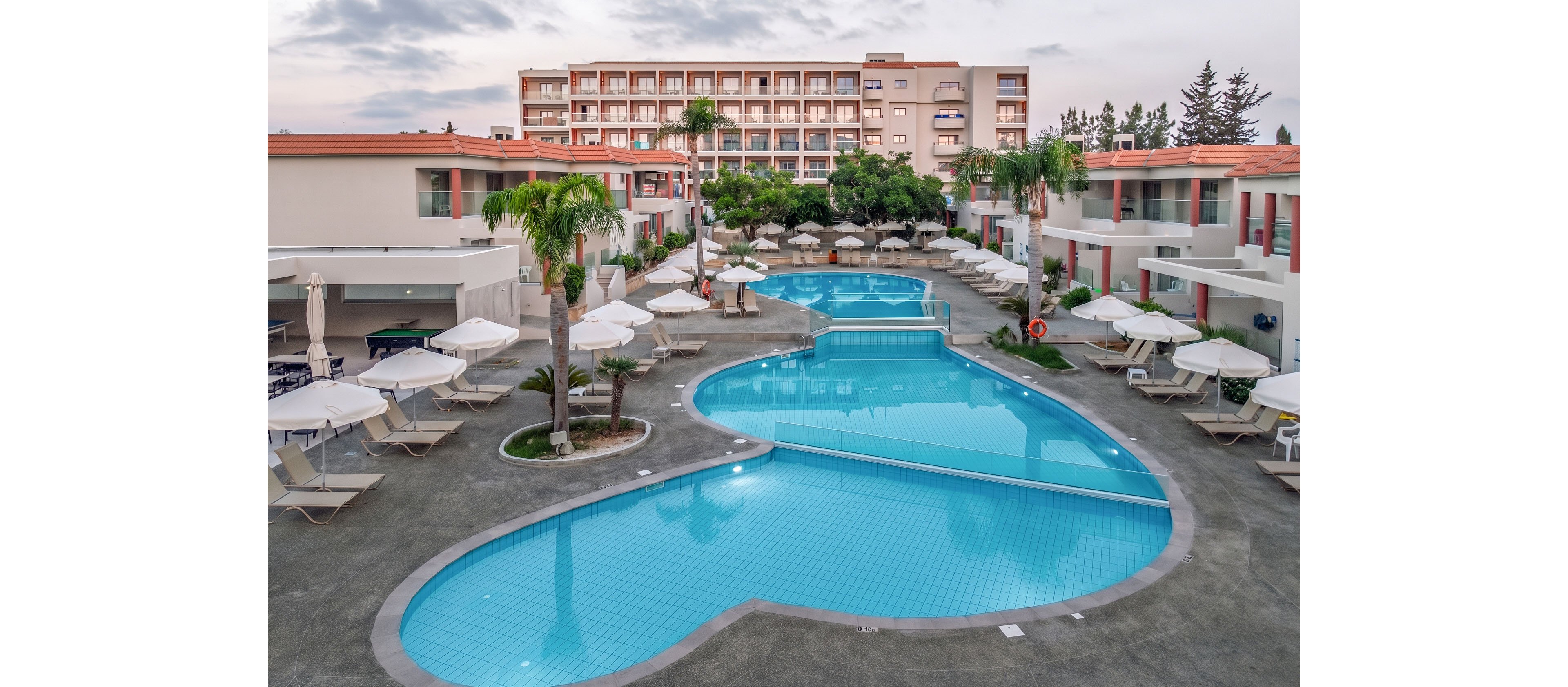 A large resort complex features multiple interconnected swimming pools surrounded by white sun loungers and umbrellas. Low-rise buildings with balconies frame the pool area, while a taller hotel structure rises in the background. Palm trees and greenery add a tropical touch to the modern courtyard setting. The soft evening light creates a calm, inviting atmosphere for vacation relaxation.