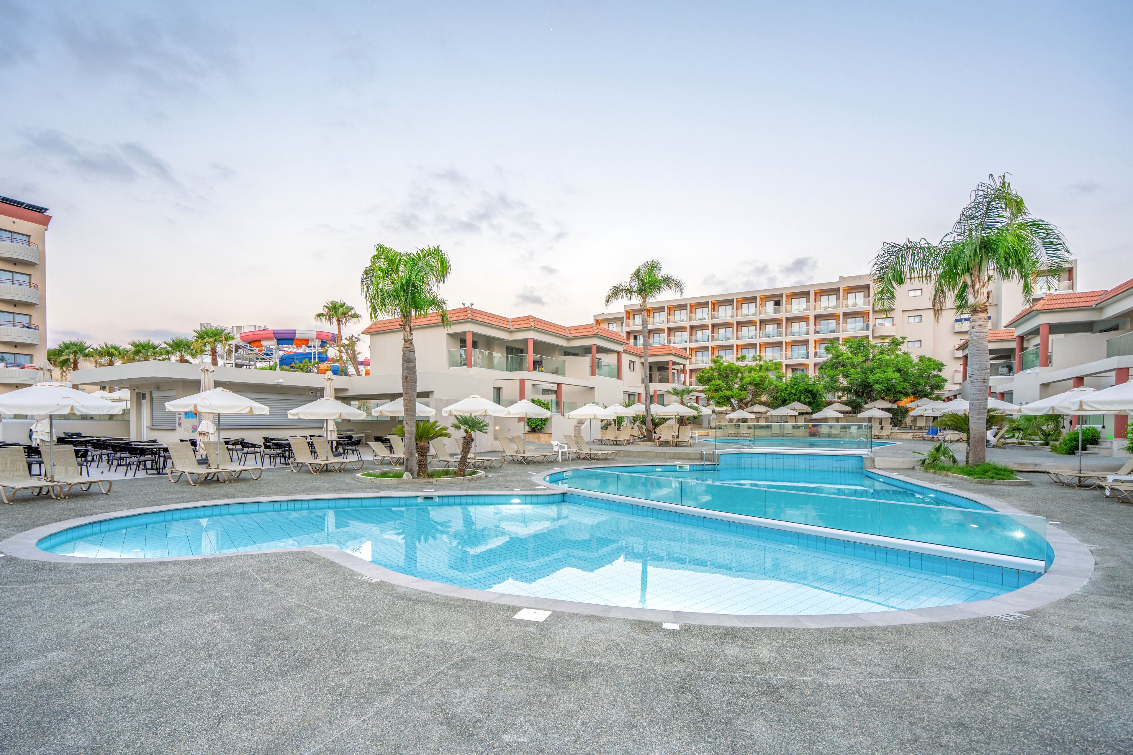 A large resort swimming pool curves through an open courtyard surrounded by sun loungers and white umbrellas. Multi-story hotel buildings frame the scene, with balconies overlooking the water and landscaped pathways. Tall palm trees and trimmed greenery add a tropical feel, while the soft early morning light creates a calm atmosphere. The clear blue sky and reflections in the pool emphasize a relaxing vacation setting.