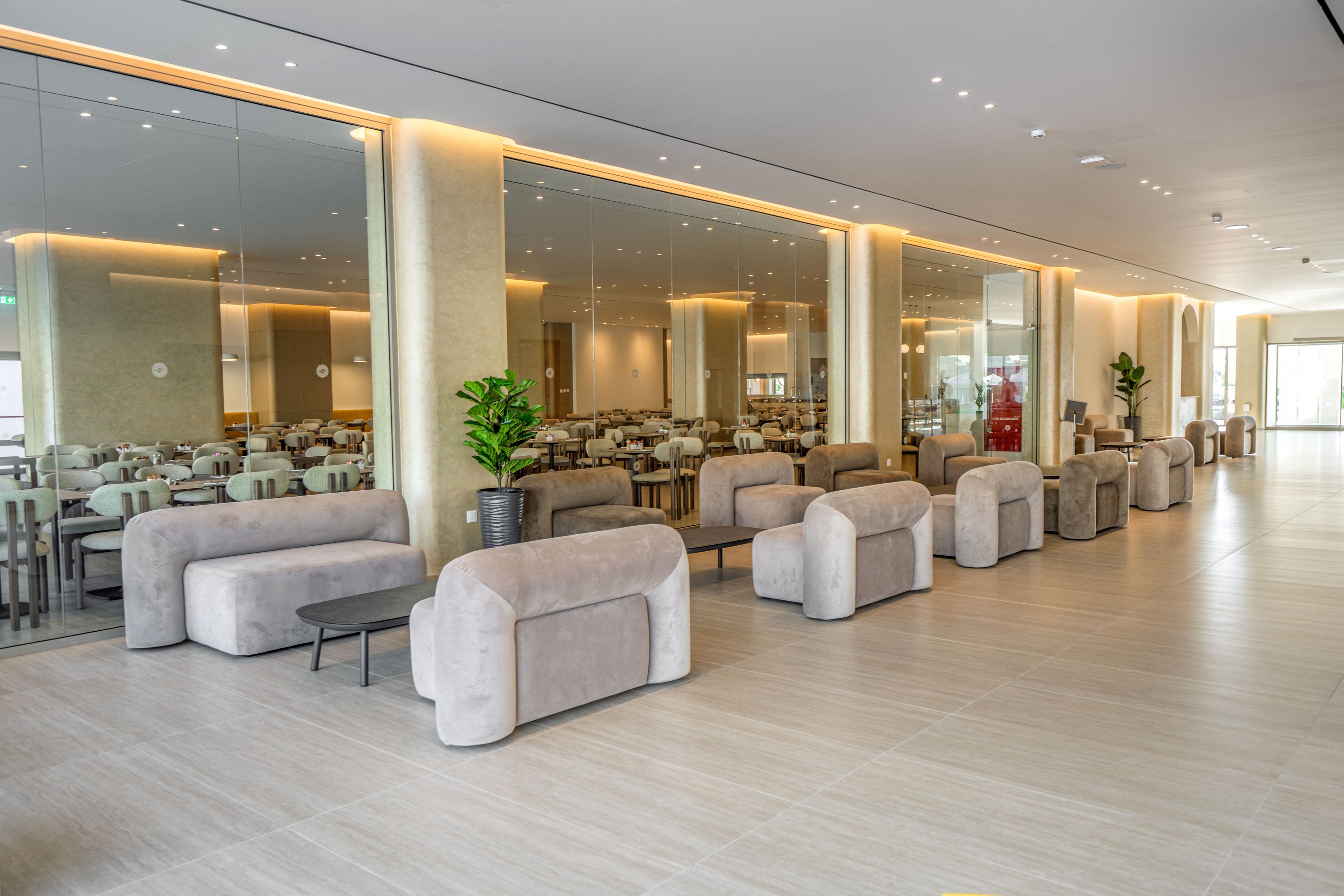 A contemporary lobby features rows of plush, light gray lounge chairs and sofas arranged along a wide corridor. Large floor-to-ceiling mirrors on the left reflect a spacious dining area with neatly aligned tables and chairs. Warm recessed lighting runs along the ceiling and wall edges, creating a soft, inviting glow. Potted green plants add a touch of color to the otherwise neutral palette, and the clean tiled floor enhances the minimalist, modern style.