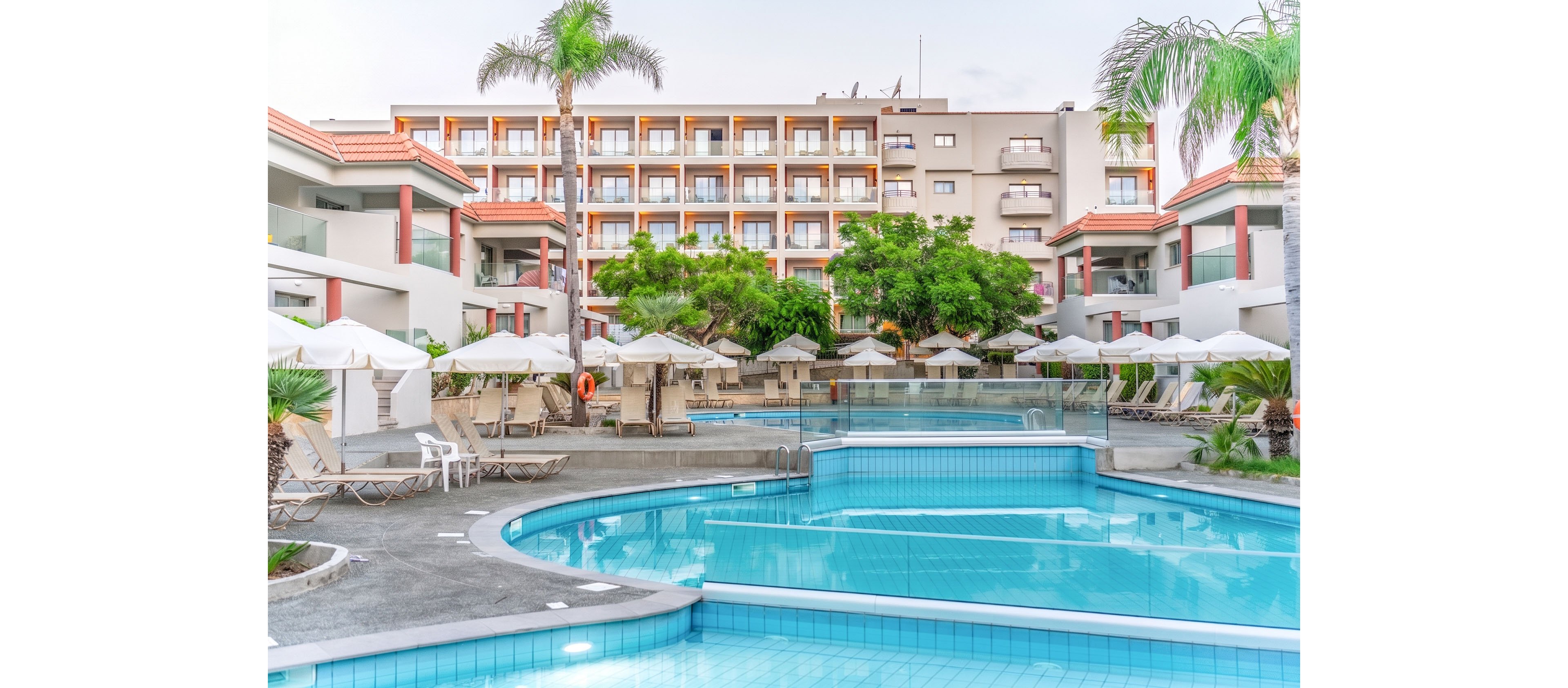 A contemporary resort hotel features a large outdoor swimming pool in the foreground. Sun loungers and white umbrellas line the poolside terrace, creating a relaxing vacation atmosphere. The multi-story hotel building with balconies rises behind lush green trees. Soft daylight and pastel tones give the scene a calm, inviting mood.