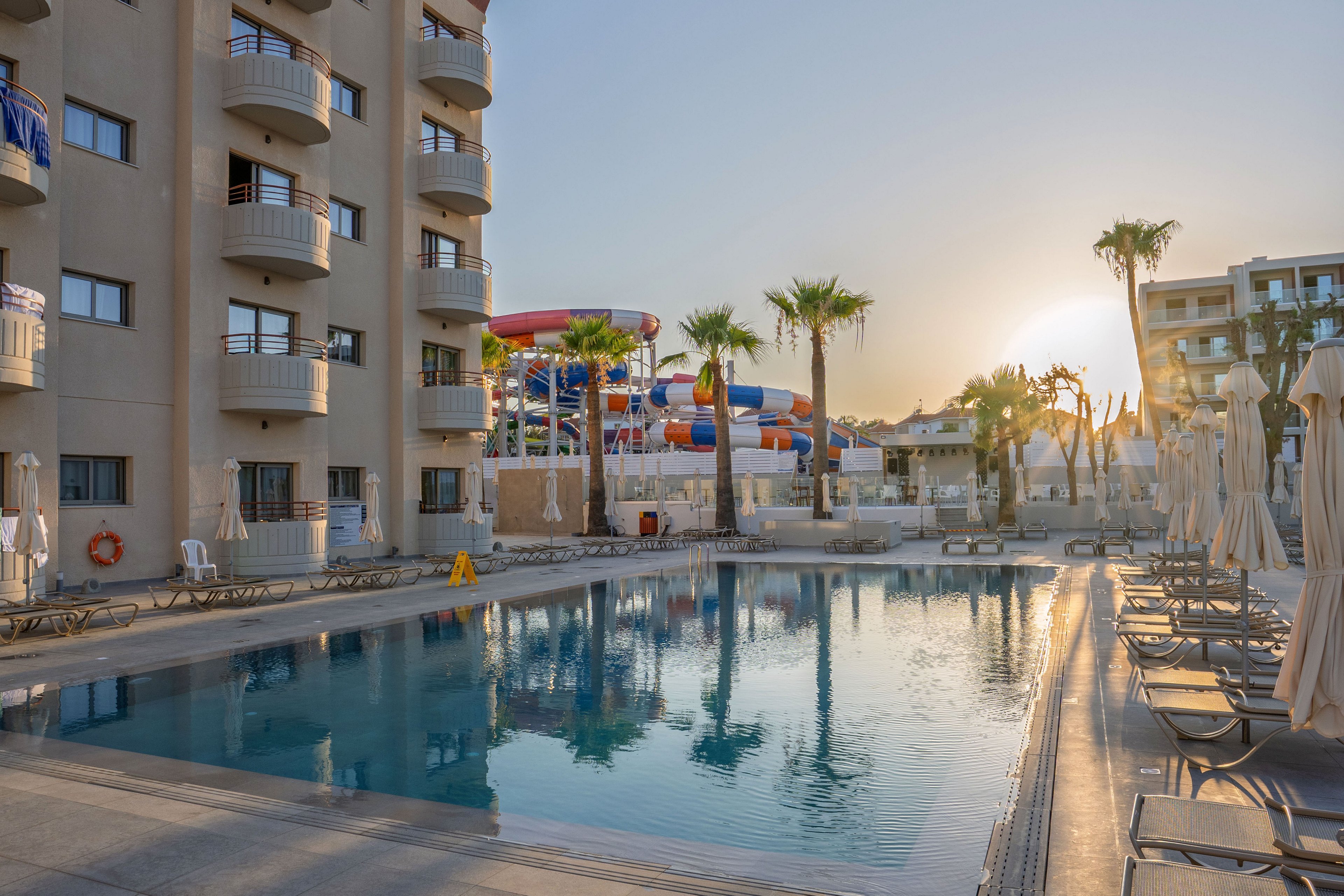 A tranquil resort swimming pool is shown at sunset, surrounded by lounge chairs and closed umbrellas. Multi-story hotel buildings with balconies frame the pool area, while tall palm trees add a tropical atmosphere. In the background, colorful water slides and structures suggest a nearby water park attraction. The calm water reflects the warm evening light, creating a relaxed vacation mood.