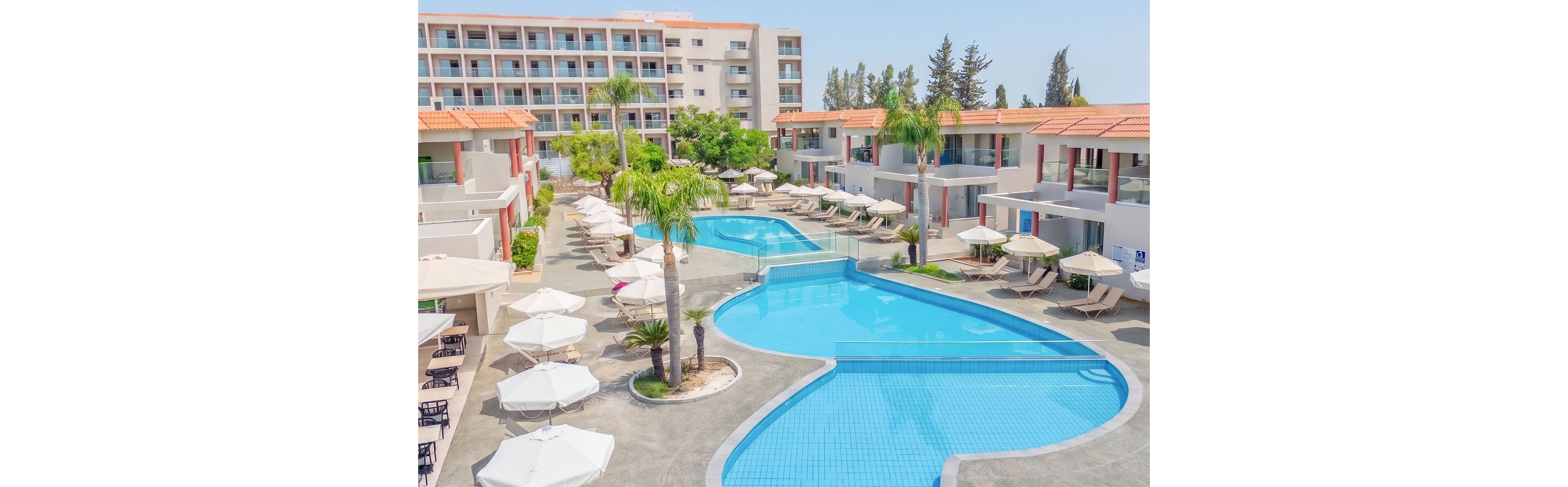 A bright resort courtyard features a large, curving outdoor swimming pool surrounded by sun loungers and white umbrellas. Modern low-rise buildings with balconies and terracotta roofs frame the pool area, creating a relaxed vacation setting. Tall palm trees and neatly arranged greenery add a tropical feel to the scene. The clear blue water and light-colored paving emphasize a clean, inviting atmosphere under sunny skies.