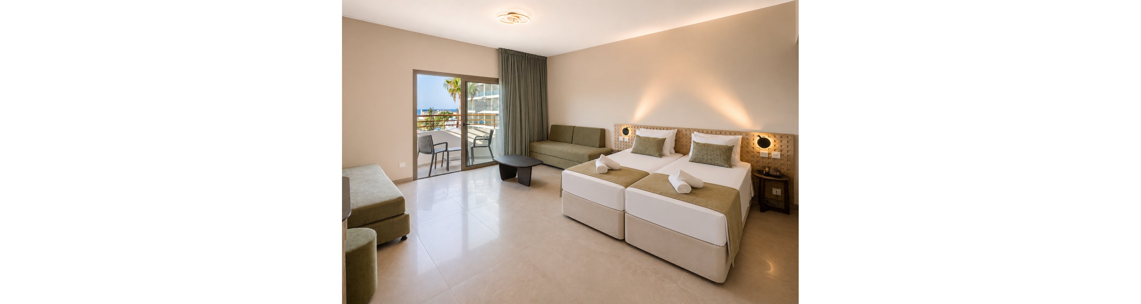 Spacious modern hotel room featuring two single beds with neatly rolled white towels and green accent cushions. A sofa and armchair sit opposite a small coffee table, creating a comfortable seating area. Large sliding glass doors open to a balcony with outdoor chairs and a partial sea view, letting in natural light. Neutral beige tones, clean lines, and minimal decor give the space a calm, contemporary atmosphere.