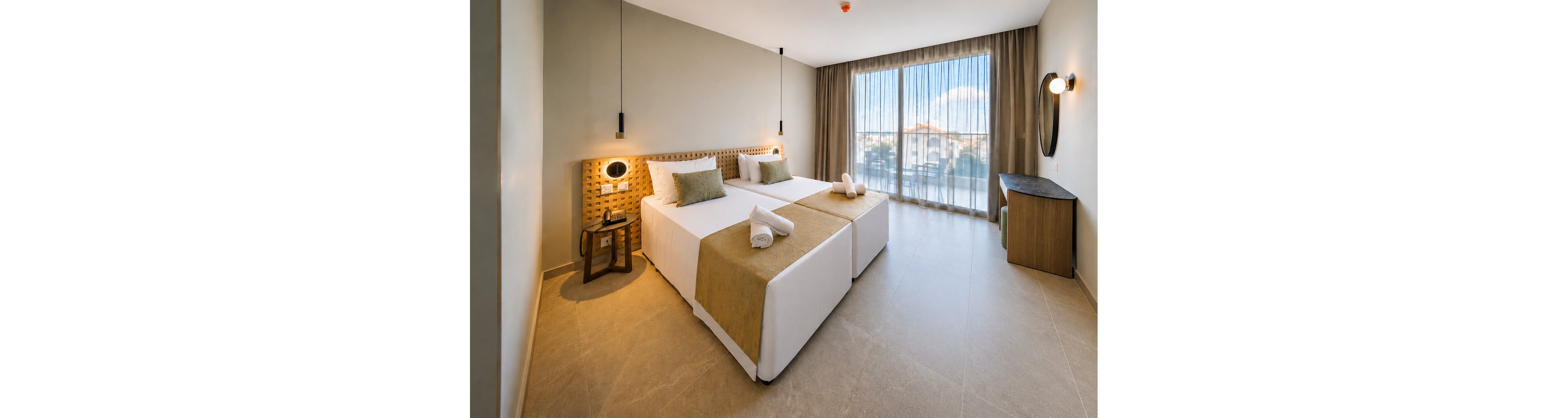 Bright contemporary hotel room featuring two neatly made twin beds with rolled white towels on top. The space includes a woven headboard, bedside table with lamp, and minimalist pendant lighting. Large floor-to-ceiling windows with sheer curtains open to a balcony and outdoor view. Neutral tones and clean lines create a calm, inviting atmosphere suitable for vacation or business travel.