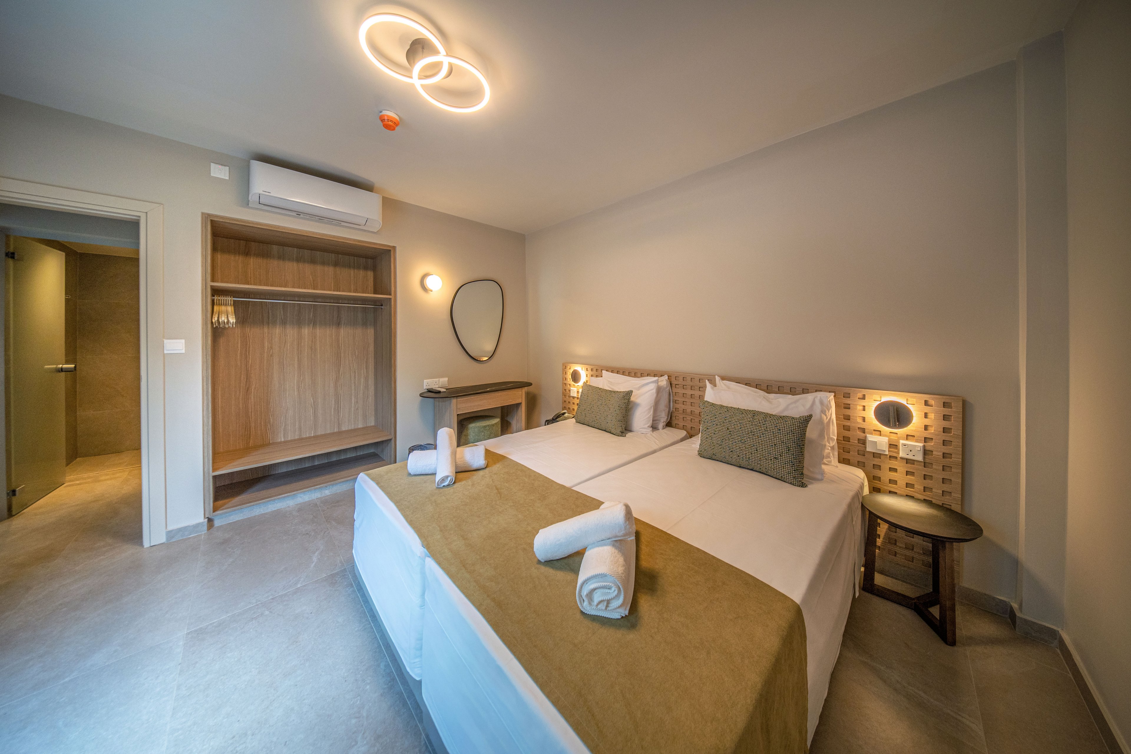 Contemporary hotel room featuring two neatly made twin beds with rolled white towels placed on top. The space includes an open wooden wardrobe, a small desk, and a round wall mirror. Soft neutral colors, warm lighting fixtures, and minimal decor create a calm, inviting atmosphere. The room appears clean, spacious, and designed for comfortable guest accommodation.