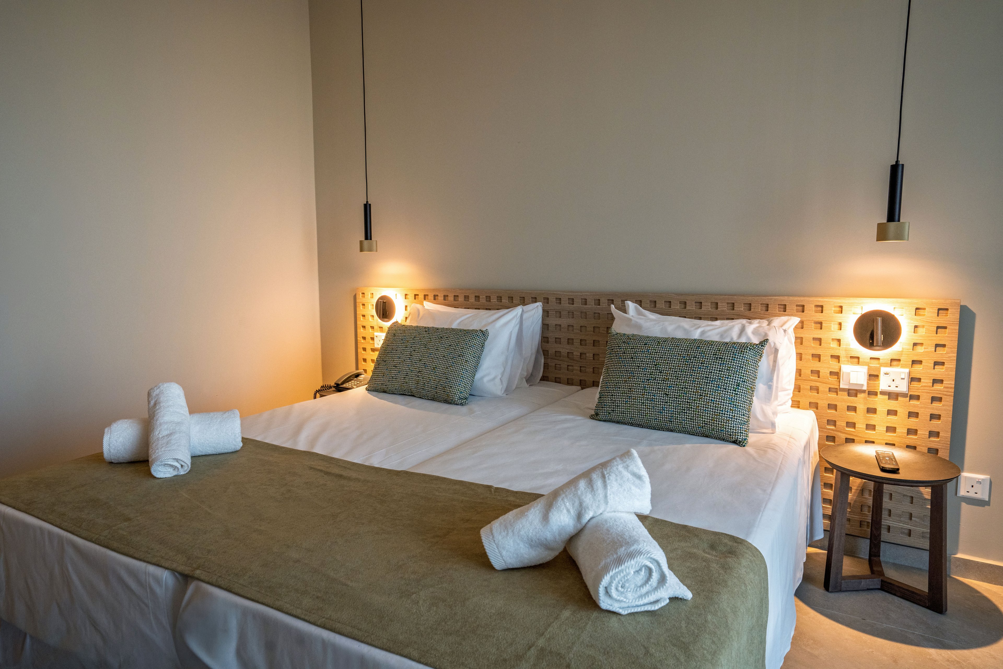 A contemporary hotel bedroom features two neatly made twin beds pushed together with white linens and a beige throw. Rolled white towels are placed on the bed, suggesting readiness for guests. Two pendant lights hang above the headboard, casting warm light and creating a cozy atmosphere. A small bedside table with a lamp and power outlet completes the minimalist, modern interior design.