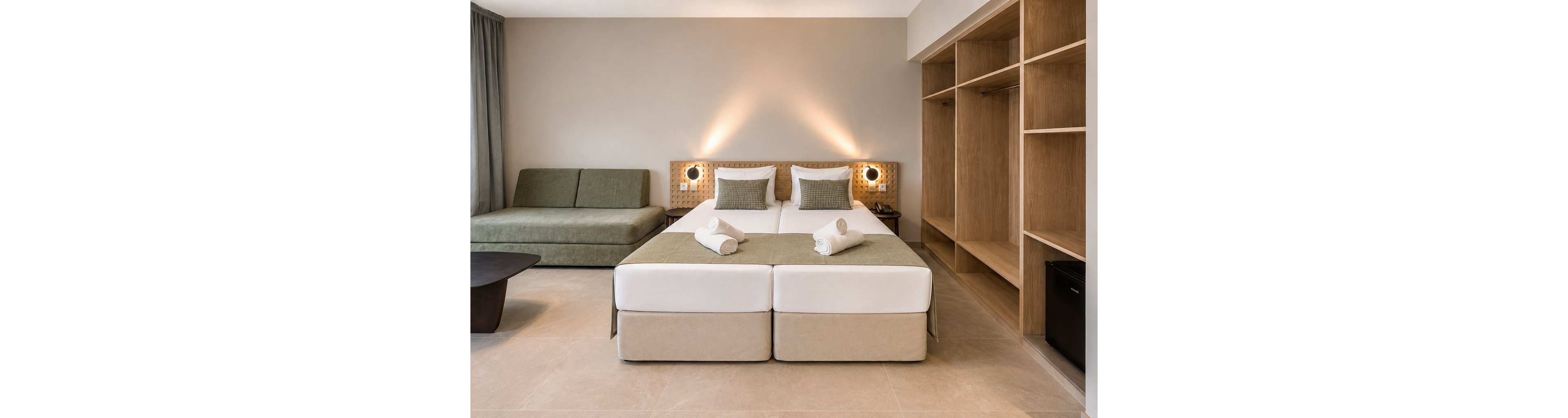 Contemporary hotel room interior featuring two neatly made twin beds with white linens and rolled towels placed on top. Soft wall-mounted lights create a warm, inviting atmosphere against a neutral beige wall. A green upholstered sofa sits to the side, complemented by an open wooden wardrobe and shelving unit. The clean lines, minimal decor, and light tiled floor emphasize a modern, comfortable design suitable for guests.