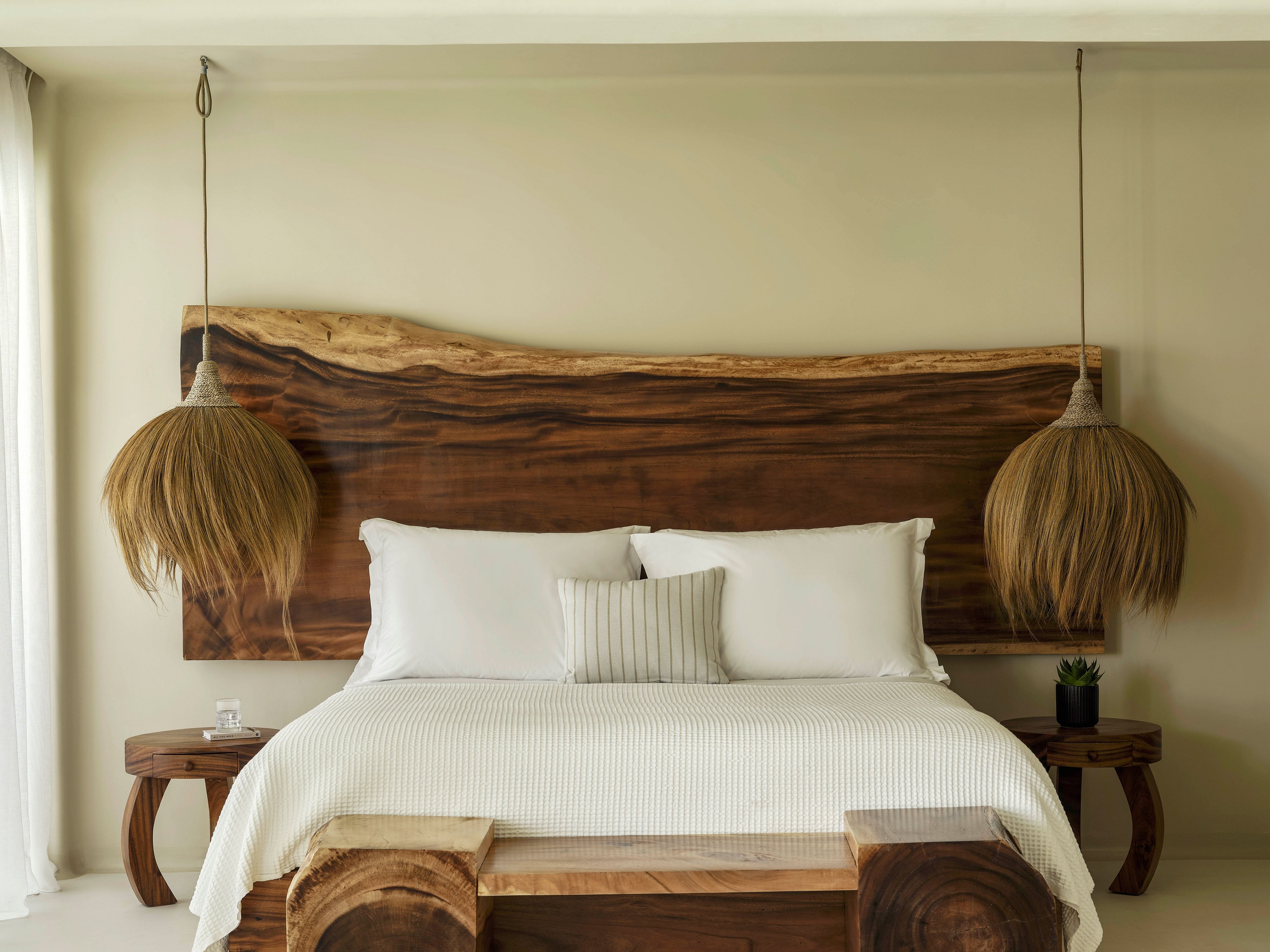 A contemporary bedroom features a large natural wood headboard behind a neatly made bed with white linens and pillows. Two hanging pendant lights with textured, straw-like shades frame the bed on either side. Matching wooden bedside tables and benches complement the organic design. Soft neutral walls and filtered daylight from a window create a calm, minimalist atmosphere.