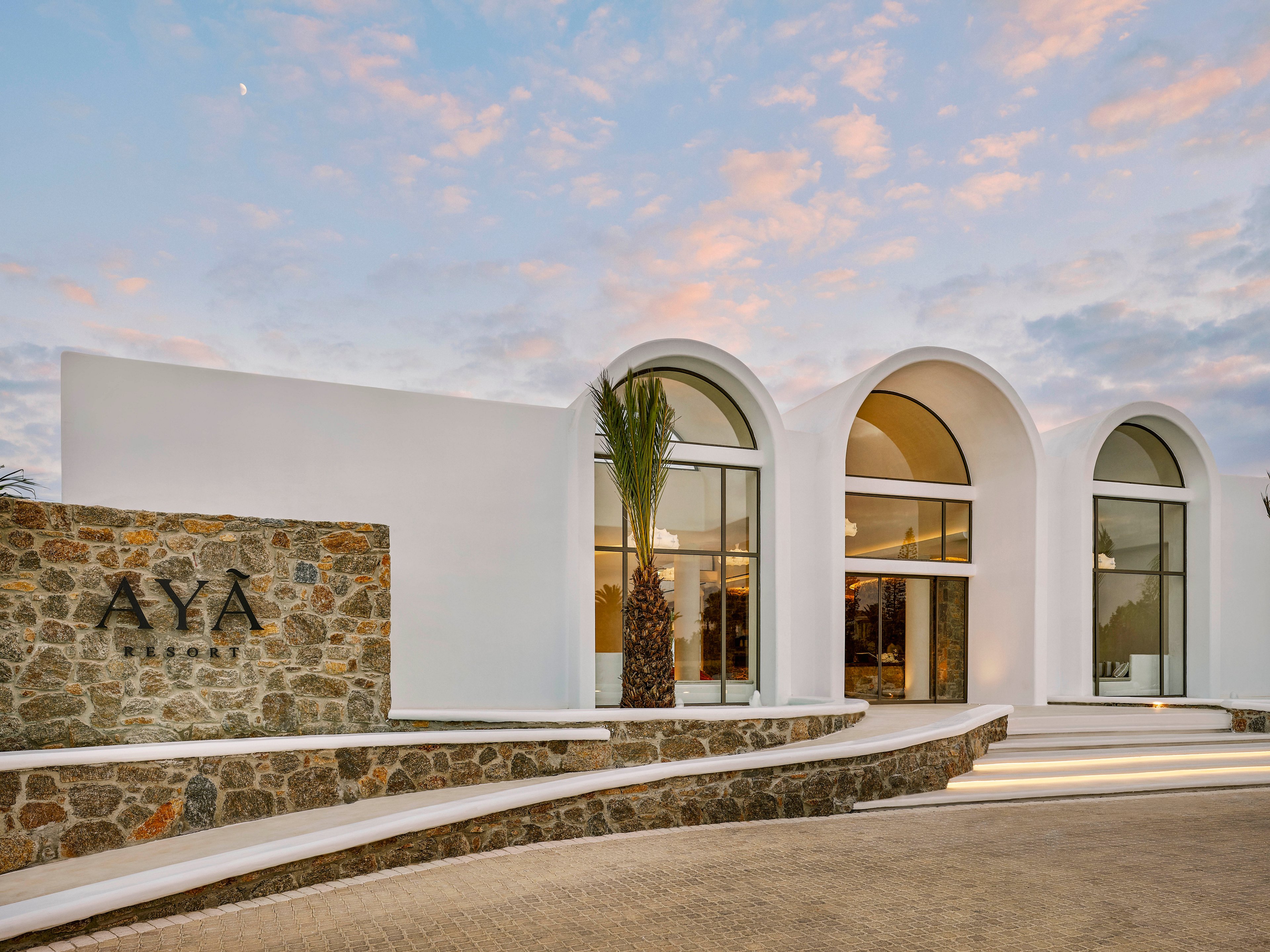 Contemporary resort architecture features a white building with large arched windows and a minimalist facade. The entrance area includes a stone wall with the clearly visible text 'AYA RESORT' and a single palm tree accenting the design. Warm interior lighting contrasts with the soft pastel colors of the evening sky. Curved pathways and clean lines create a luxurious, inviting atmosphere.