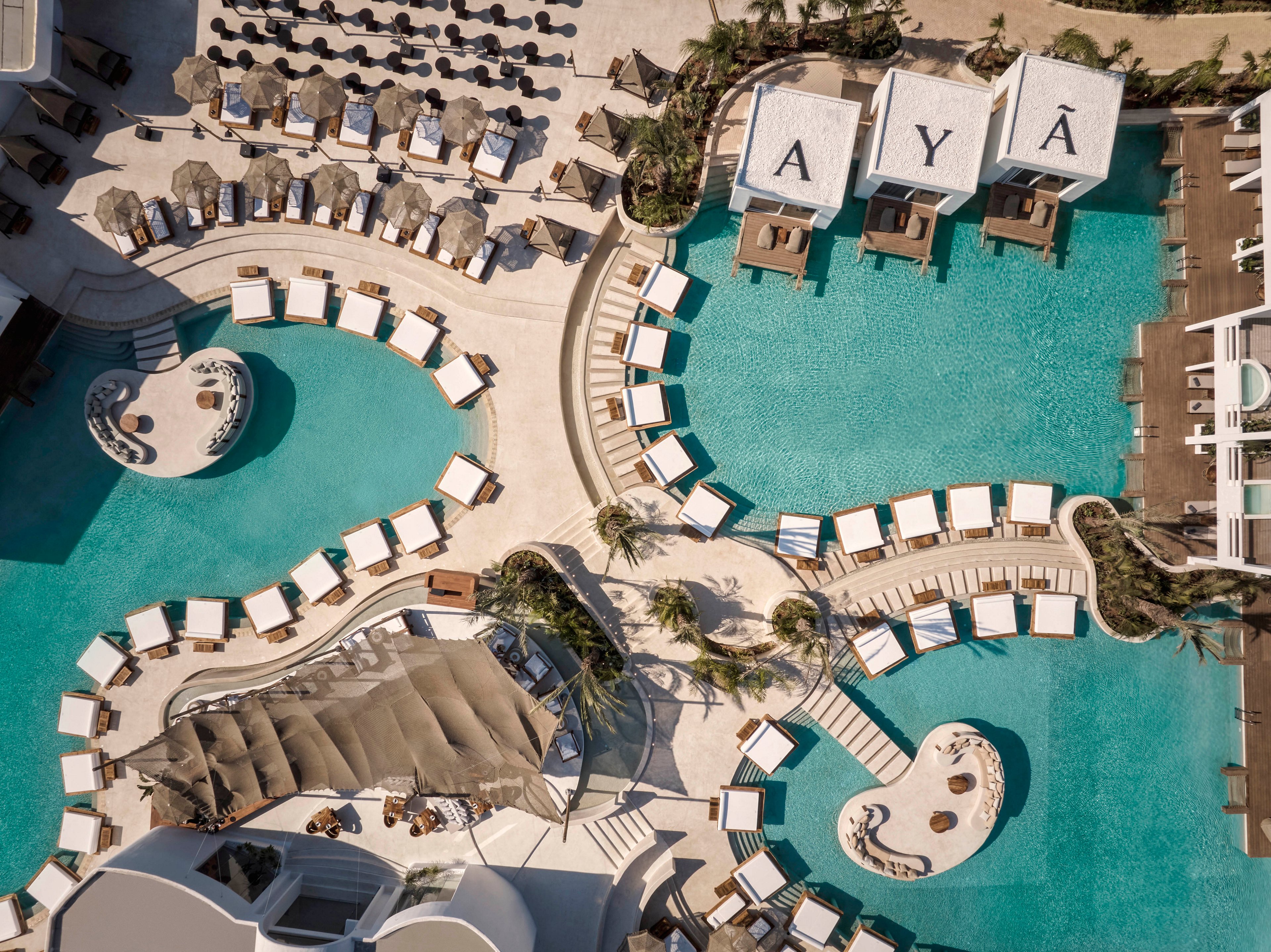 Aerial photograph of a luxury resort featuring multiple turquoise swimming pools and curved sun deck areas. Numerous lounge chairs and umbrellas are arranged neatly around the pools, creating an inviting vacation atmosphere. Three cabanas with the letters A, Y, and Ã visible on their roofs overlook one of the larger pools. Palm trees, shaded seating areas, and organic-shaped islands in the water add to the tropical, upscale design.