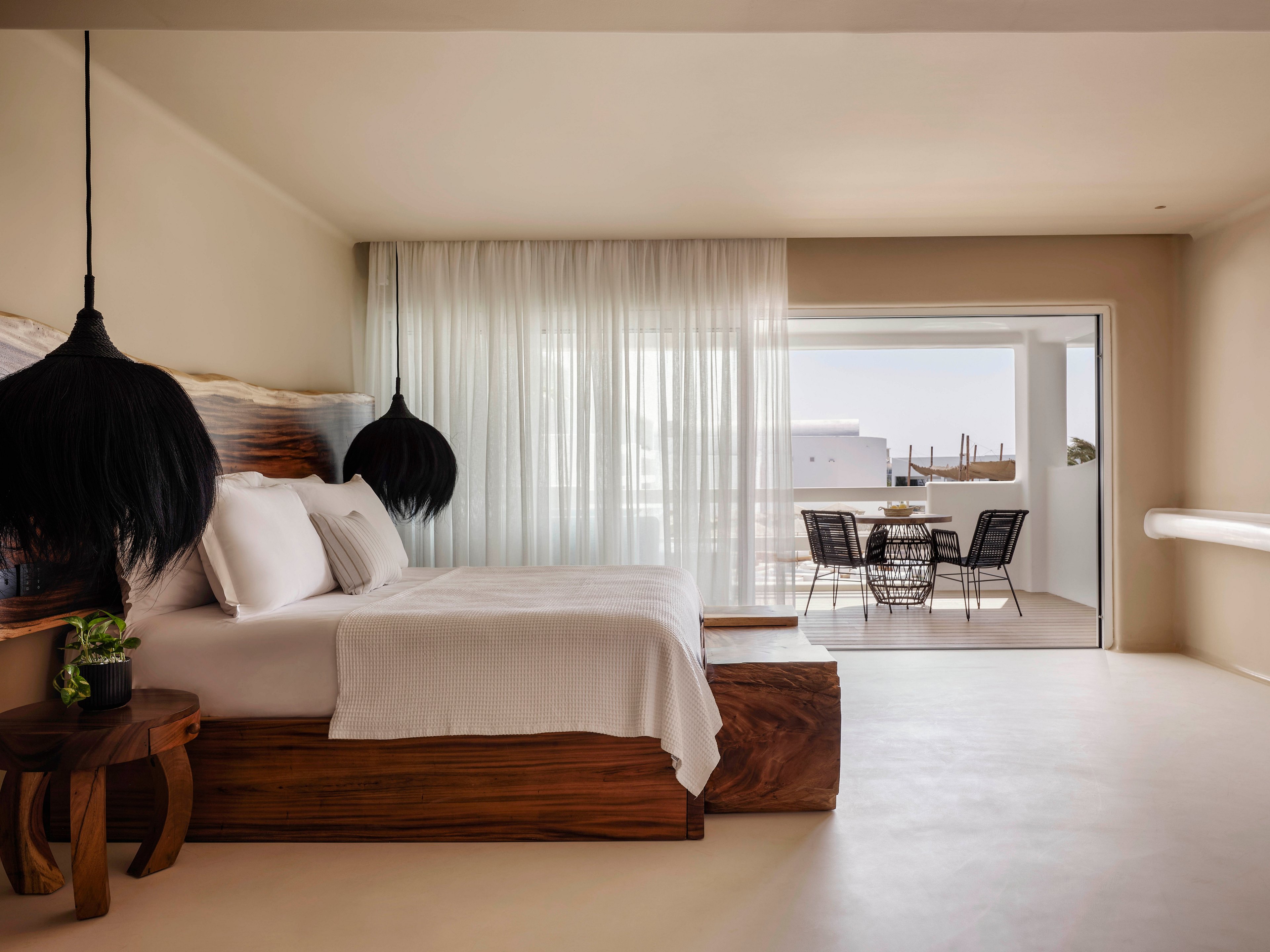 A modern minimalist hotel bedroom features a large wooden bed with crisp white linens and pillows. Sheer white curtains cover the glass doors leading to a bright balcony. Black pendant lights and a small wooden side table with a potted plant add contrast to the neutral interior. Outside, a table with four chairs overlooks a sunlit terrace and distant buildings.