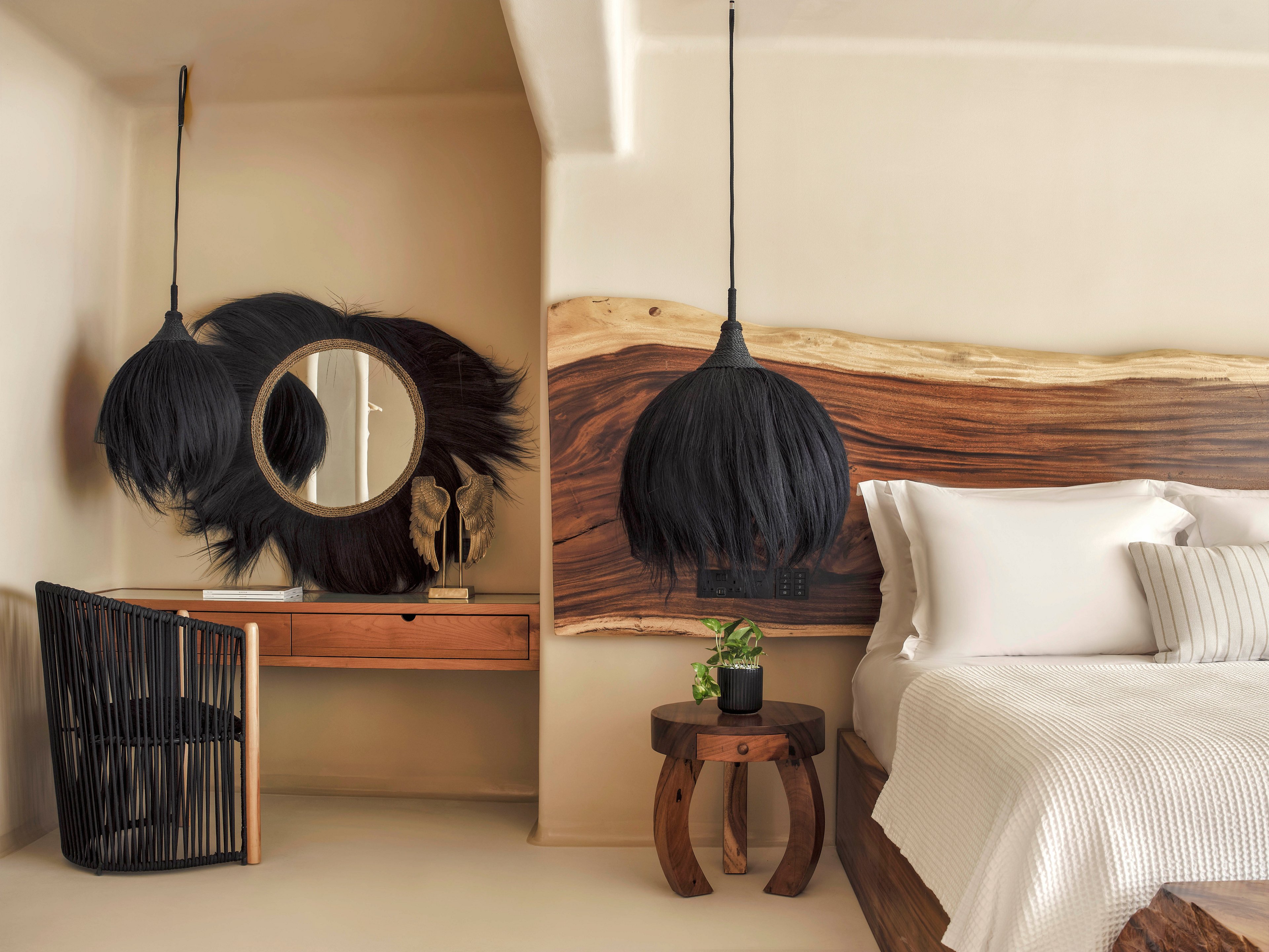 A contemporary bedroom features a large bed with white linens against a wall accented by a live-edge wooden headboard. Sculptural black pendant lights with feathered textures hang above both the bedside table and the adjacent vanity desk. A round mirror framed by black decorative elements and a woven black chair add contrast to the neutral beige walls and floor. The overall design blends natural wood with modern bohemian decor for a warm, stylish retreat.