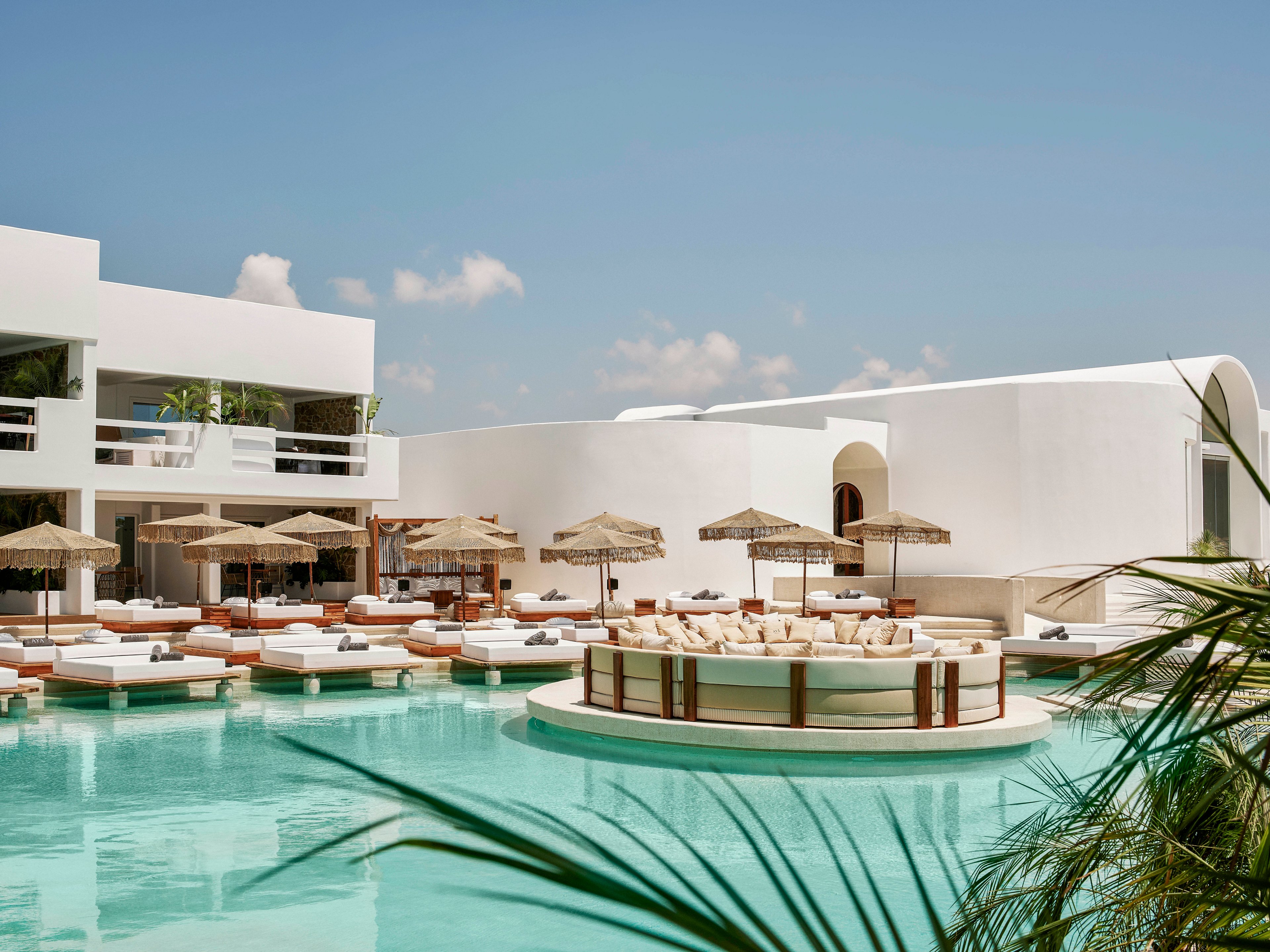 A contemporary white resort features a large turquoise swimming pool surrounded by cushioned sun loungers and parasols. The central circular island in the pool offers additional seating, creating a social focal point. Minimalist architecture with clean lines and balconies frames the scene under a bright blue sky with scattered clouds. Tropical plants in the foreground add a touch of greenery to the serene, upscale atmosphere.