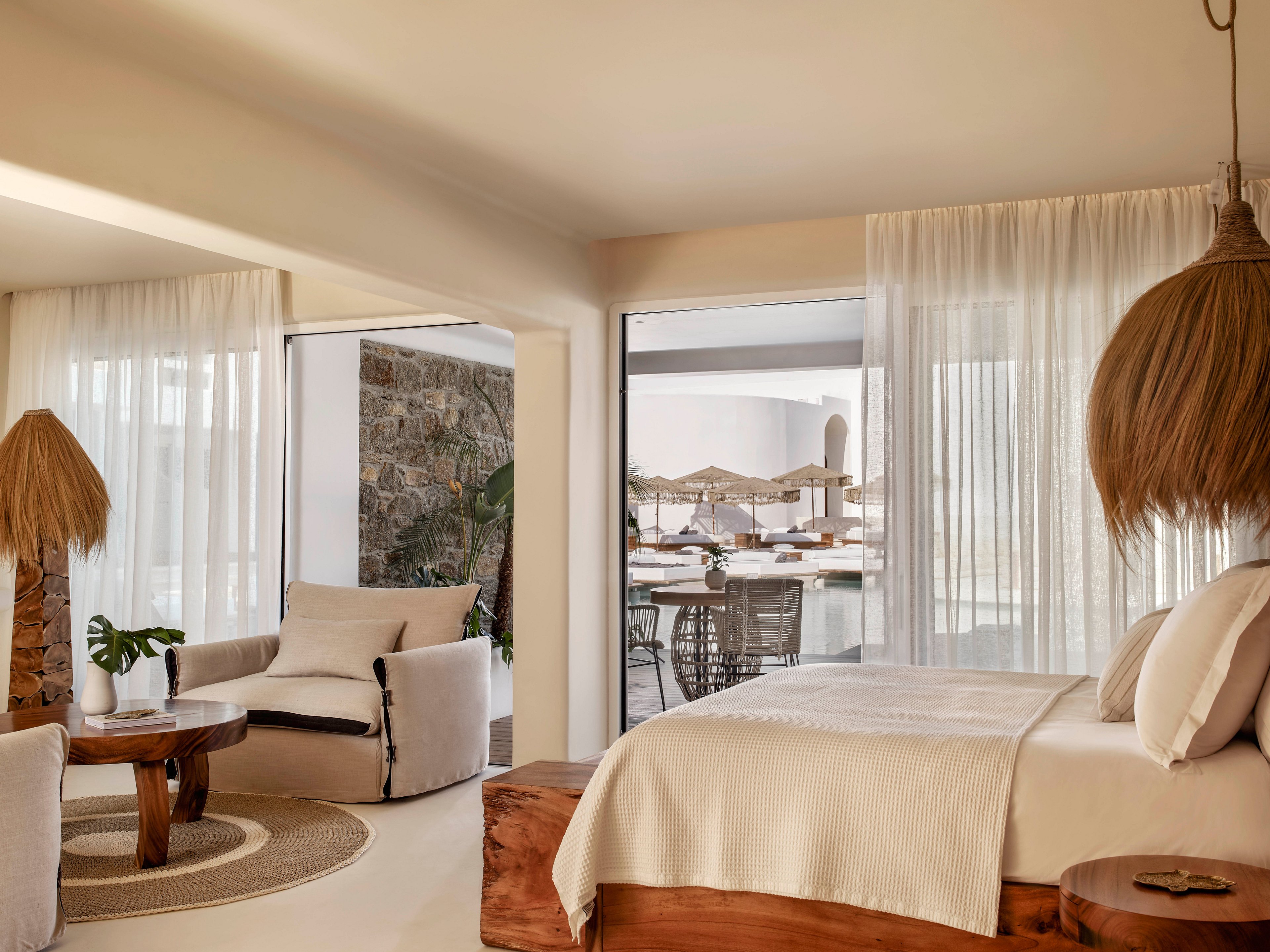 A bright, contemporary bedroom features a large bed with white linens and warm wooden furniture. Floor-to-ceiling sheer curtains frame sliding glass doors that open to a terrace with outdoor seating and umbrellas. Natural textures such as woven lampshades, a round jute rug, and potted greenery create a relaxed, organic atmosphere. Soft neutral tones and abundant daylight give the space a serene, coastal resort feel.