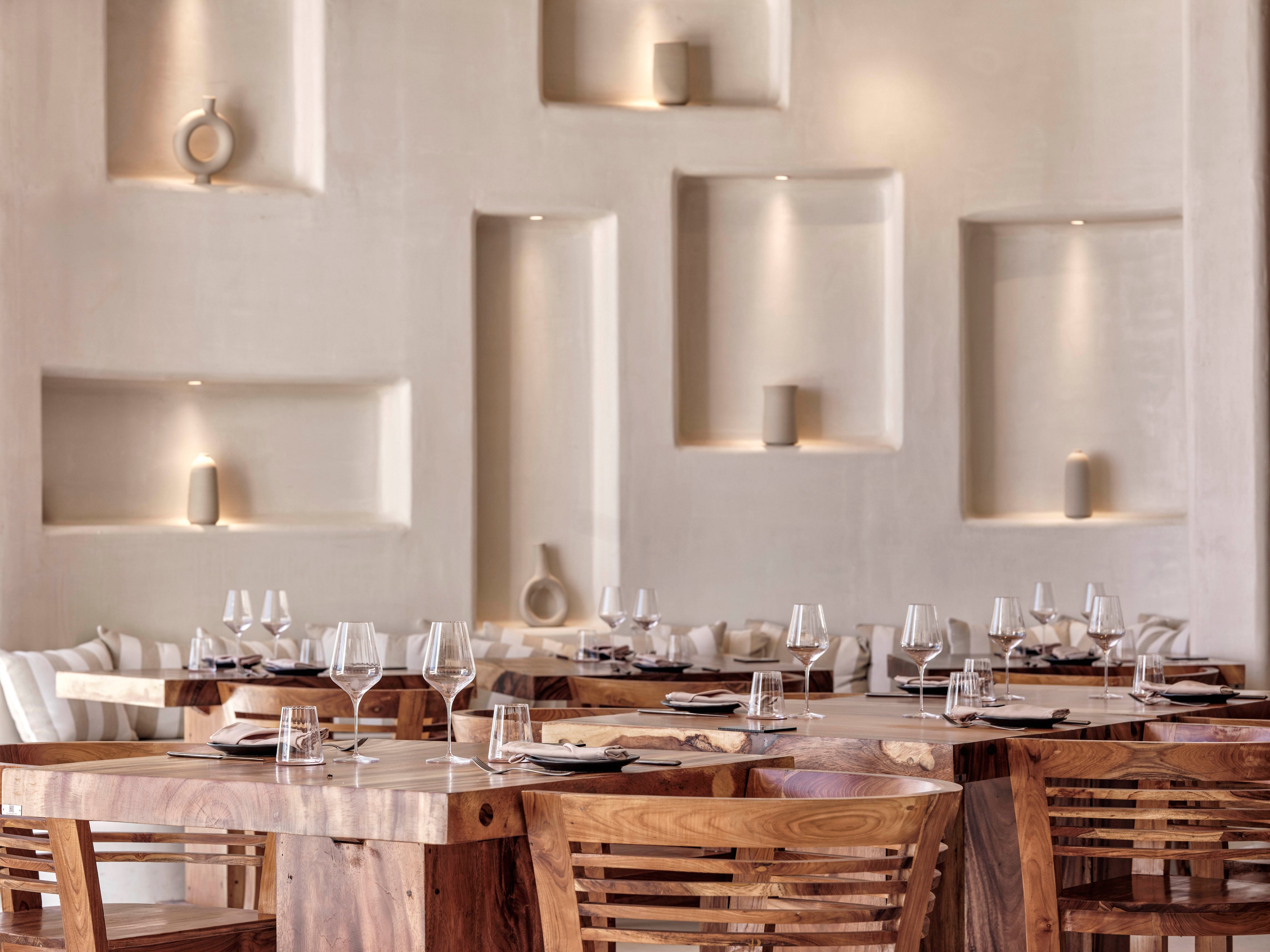 A contemporary restaurant interior features solid wooden tables and slatted wooden chairs arranged neatly in rows. Each table is set with clear wine glasses, water glasses, and dark plates, suggesting a fine dining experience. The light-colored wall behind the seating area has recessed niches with simple decorative objects and subtle built-in lighting. The overall design is minimalist and warm, combining natural wood tones with soft neutral walls for an inviting atmosphere.