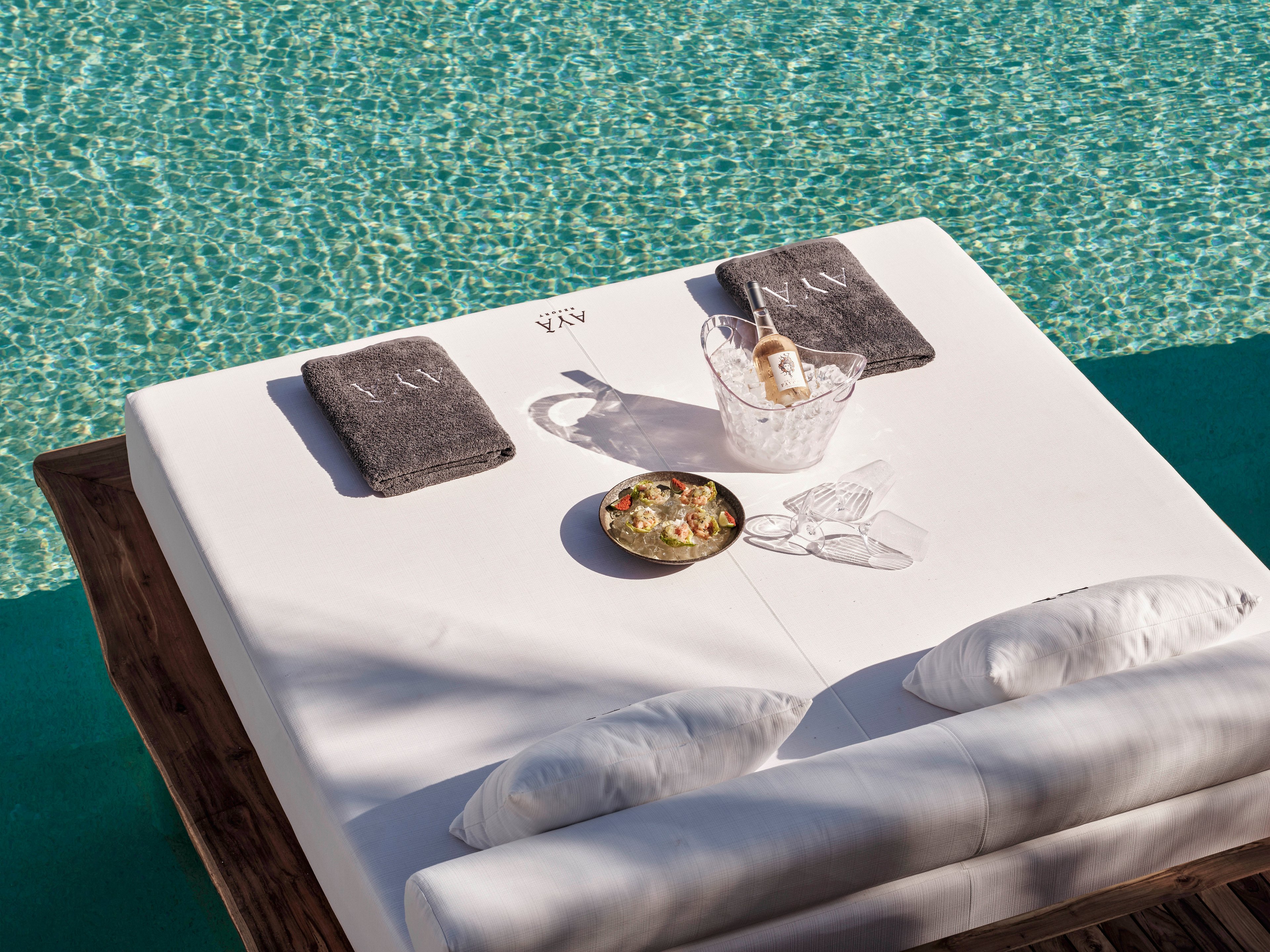 A white daybed is set beside a clear turquoise swimming pool, creating a relaxed resort atmosphere. On the bed, two gray folded towels and two white pillows are neatly arranged. In the center, a bowl of food and a bucket with a drink and ice suggest a casual poolside snack. The bright sunlight and clean lines emphasize a calm, luxurious vacation setting.