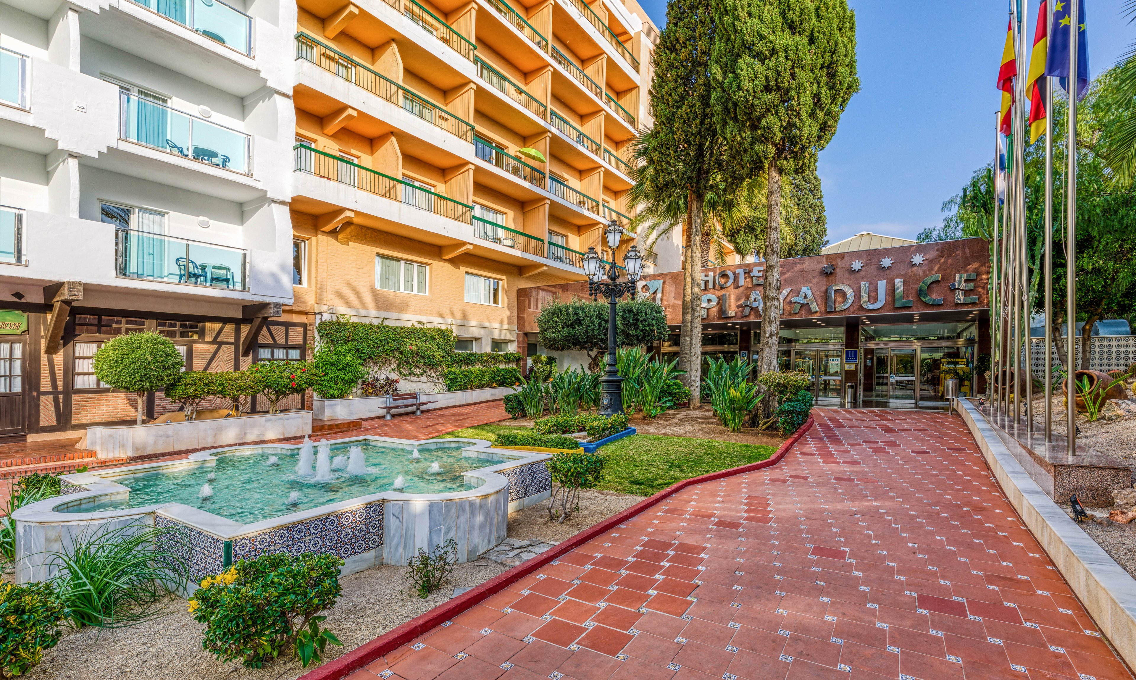 A hotel entrance is shown with a tiled walkway leading toward glass doors. On the left, a decorative fountain with clear water and patterned tiles sits among landscaped greenery. A multi-story building with balconies rises above the courtyard, while tall trees and plants add a lush atmosphere. Several national flags line the right side of the path, suggesting an international tourist destination.