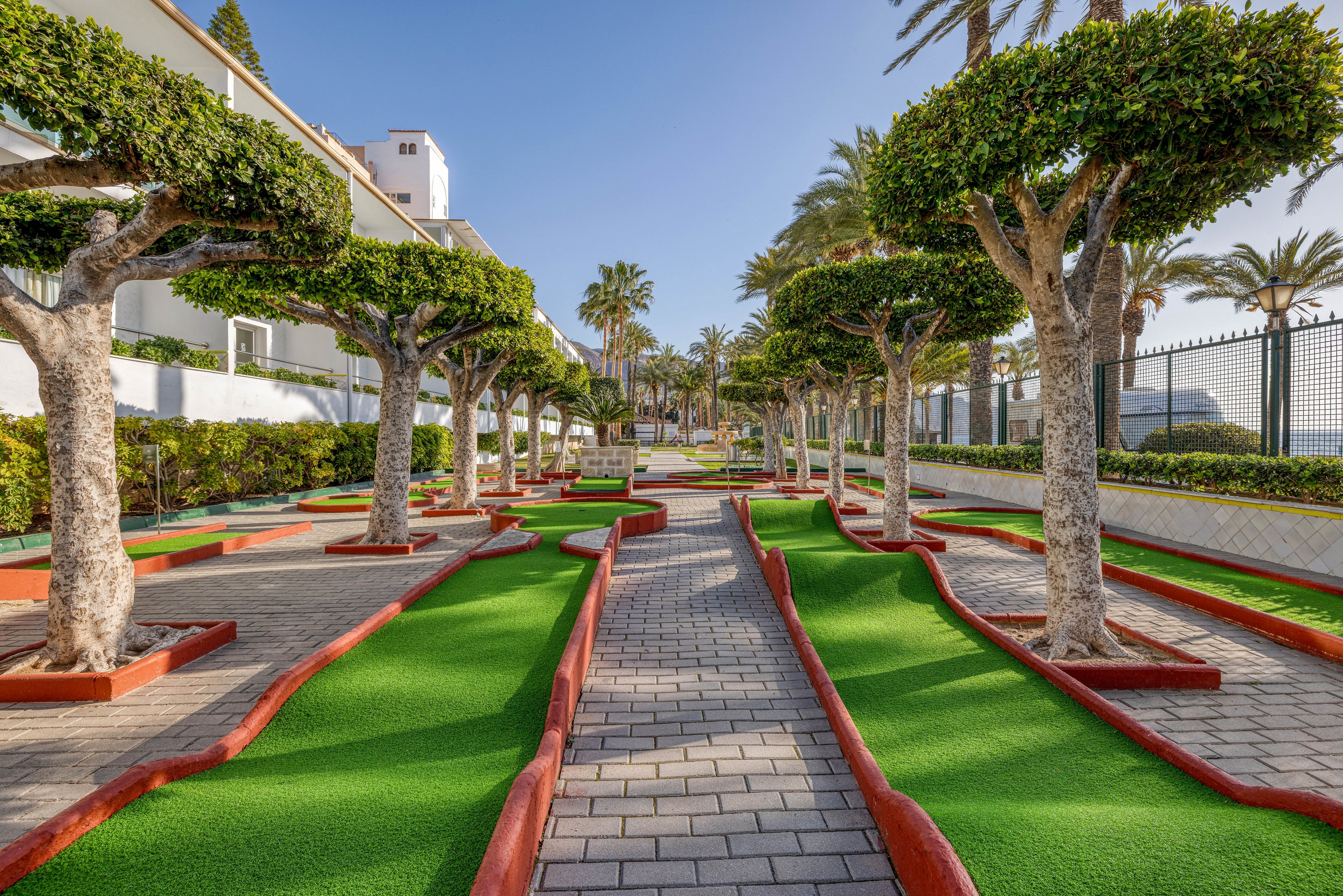 A neatly designed mini golf course lines a paved walkway between trimmed trees at a sunny resort. Bright green artificial turf contrasts with red borders and gray stone pavers. Palm trees and white buildings frame the scene, suggesting a coastal or vacation destination. The area appears quiet and well-maintained, ideal for leisurely outdoor recreation.