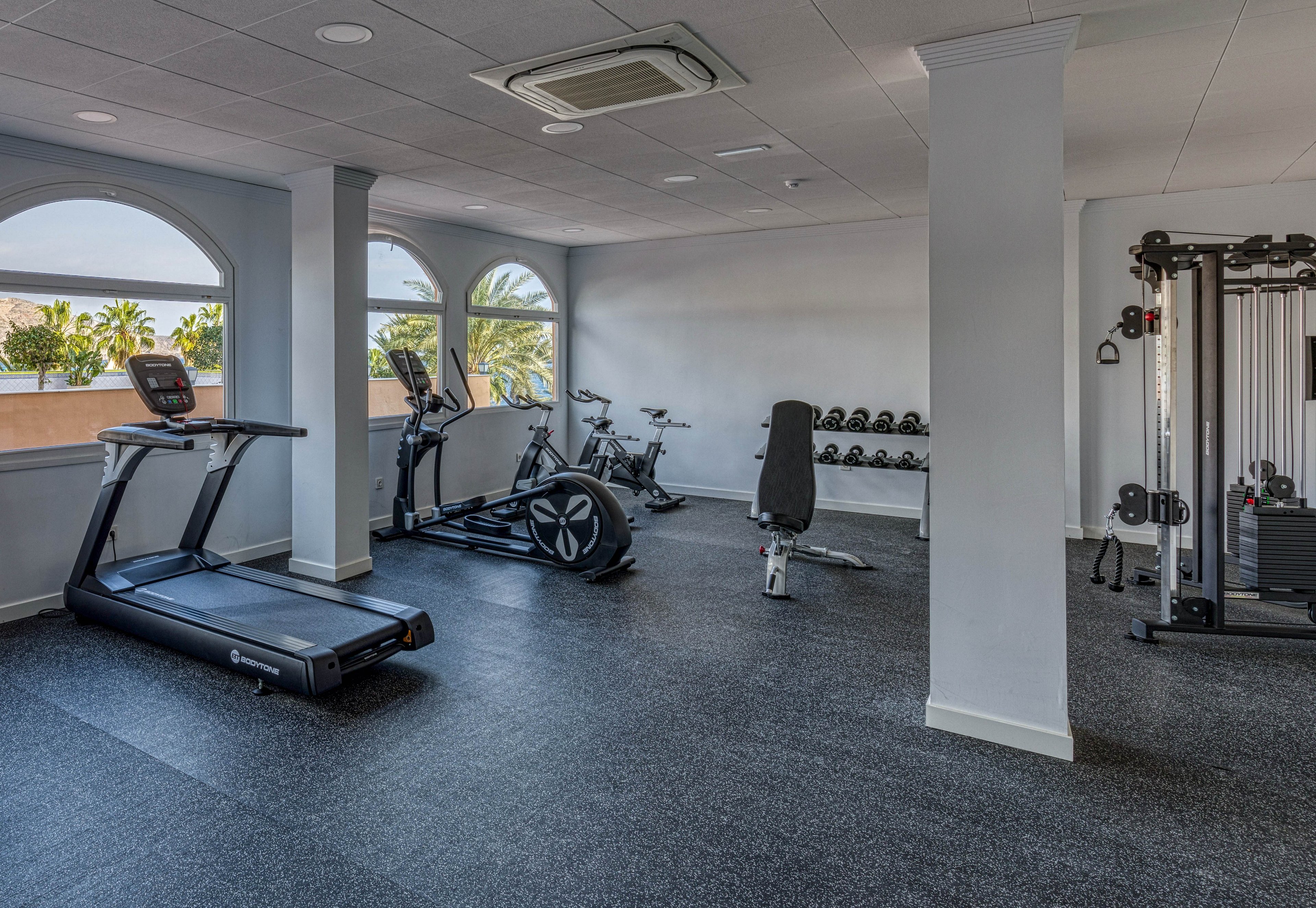 A clean, modern gym room features various exercise machines and free weights. Cardio equipment such as a treadmill and stationary bikes are arranged near large arched windows. A rack of dumbbells and a multi-station weight machine provide strength training options. Natural light fills the space, highlighting the minimalist design and spacious layout.
