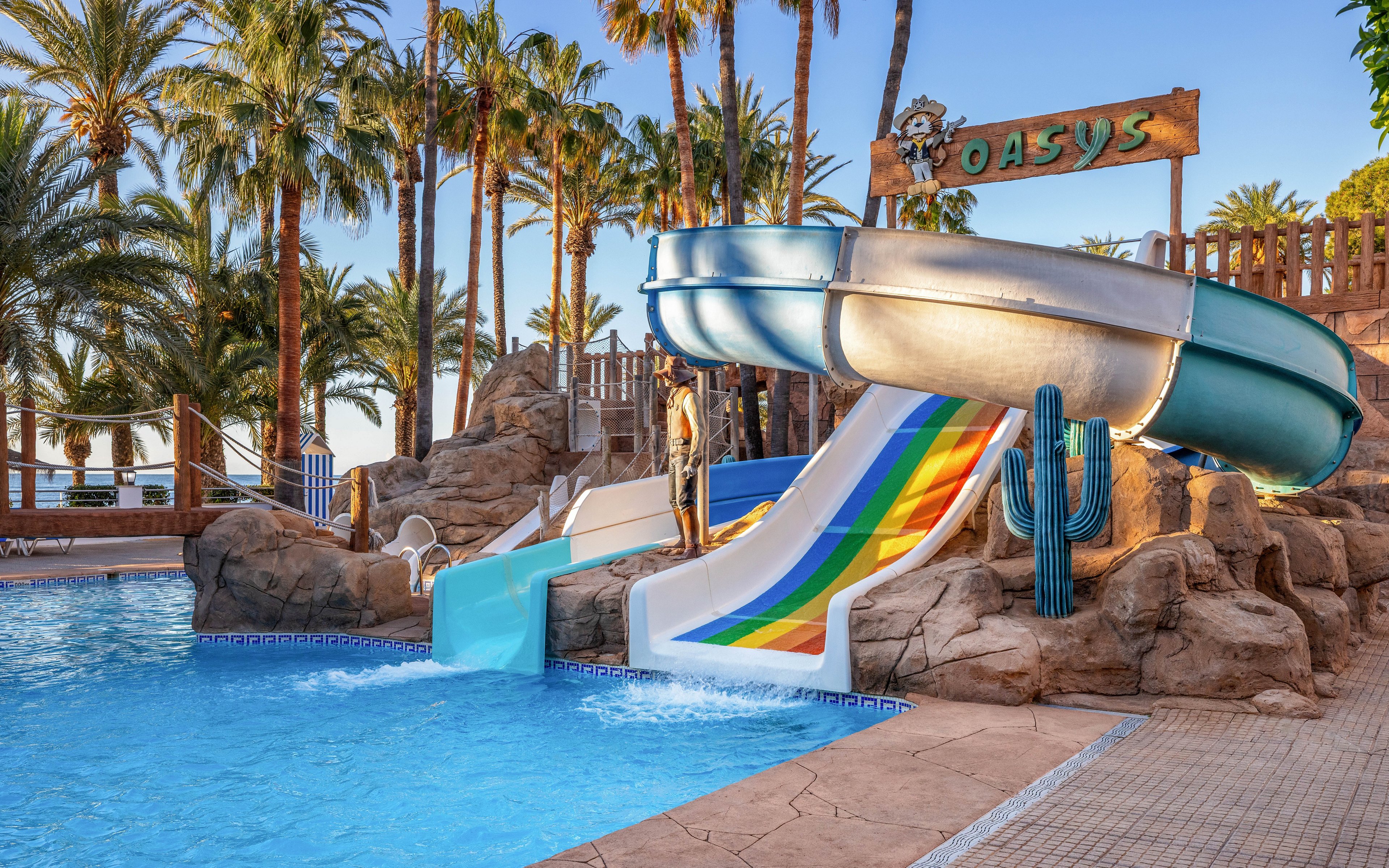 A vibrant outdoor water park features several colorful slides emptying into a clear blue swimming pool. Palm trees and rocky landscaping create a tropical oasis atmosphere around the attraction. A person stands near the top of the slides, while empty lounge chairs sit by the poolside. A wooden sign reading "OASYS" is visible above the slides, enhancing the resort-like setting.