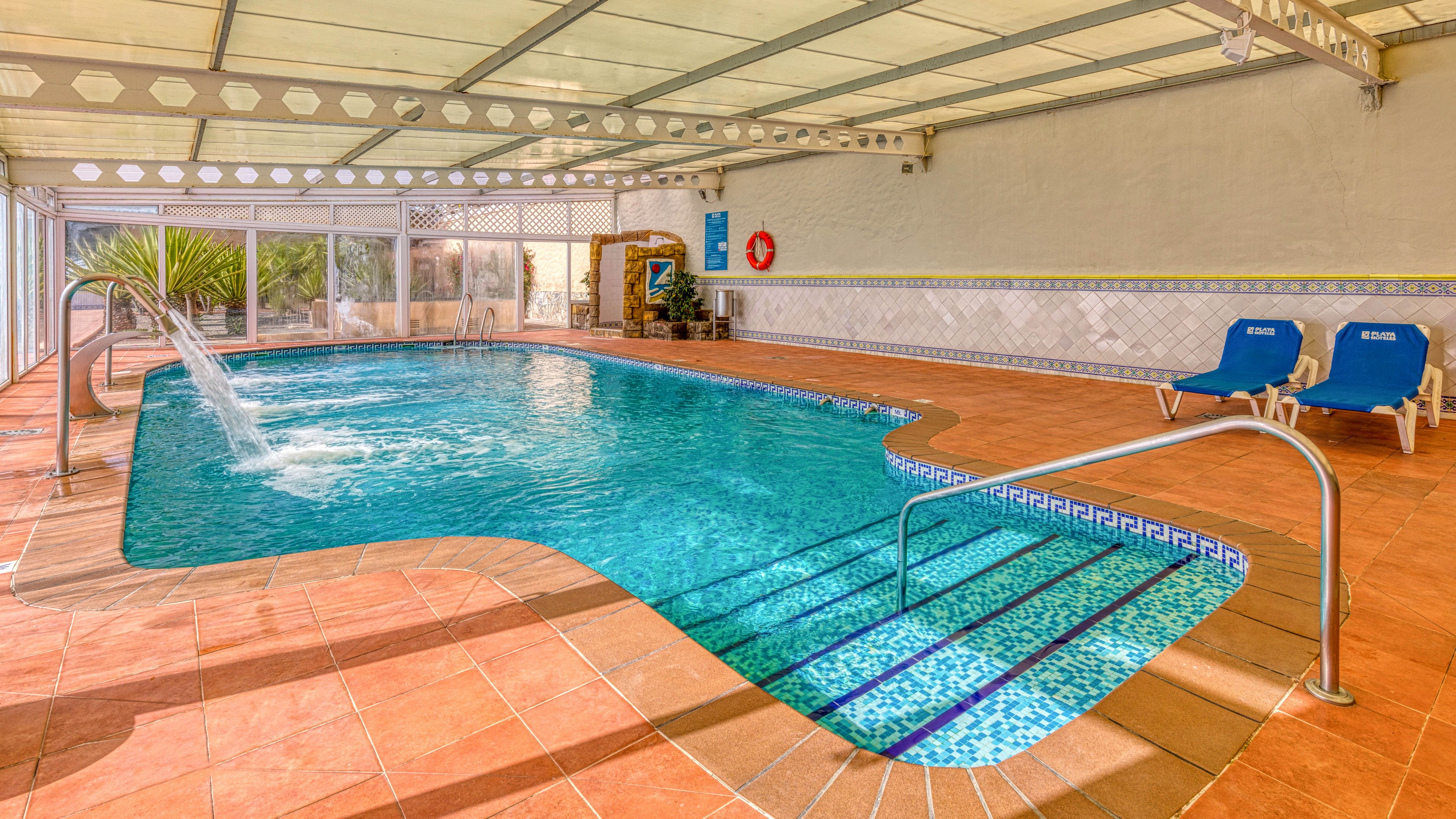 An indoor swimming pool with clear blue water is shown under a translucent roof. A curved waterfall feature pours water into the pool near the shallow end. Metal handrails lead into the water, and tiled steps are visible beneath the surface. Two blue lounge chairs sit on the terracotta-tiled deck beside the pool, with plants and safety equipment along the far wall.