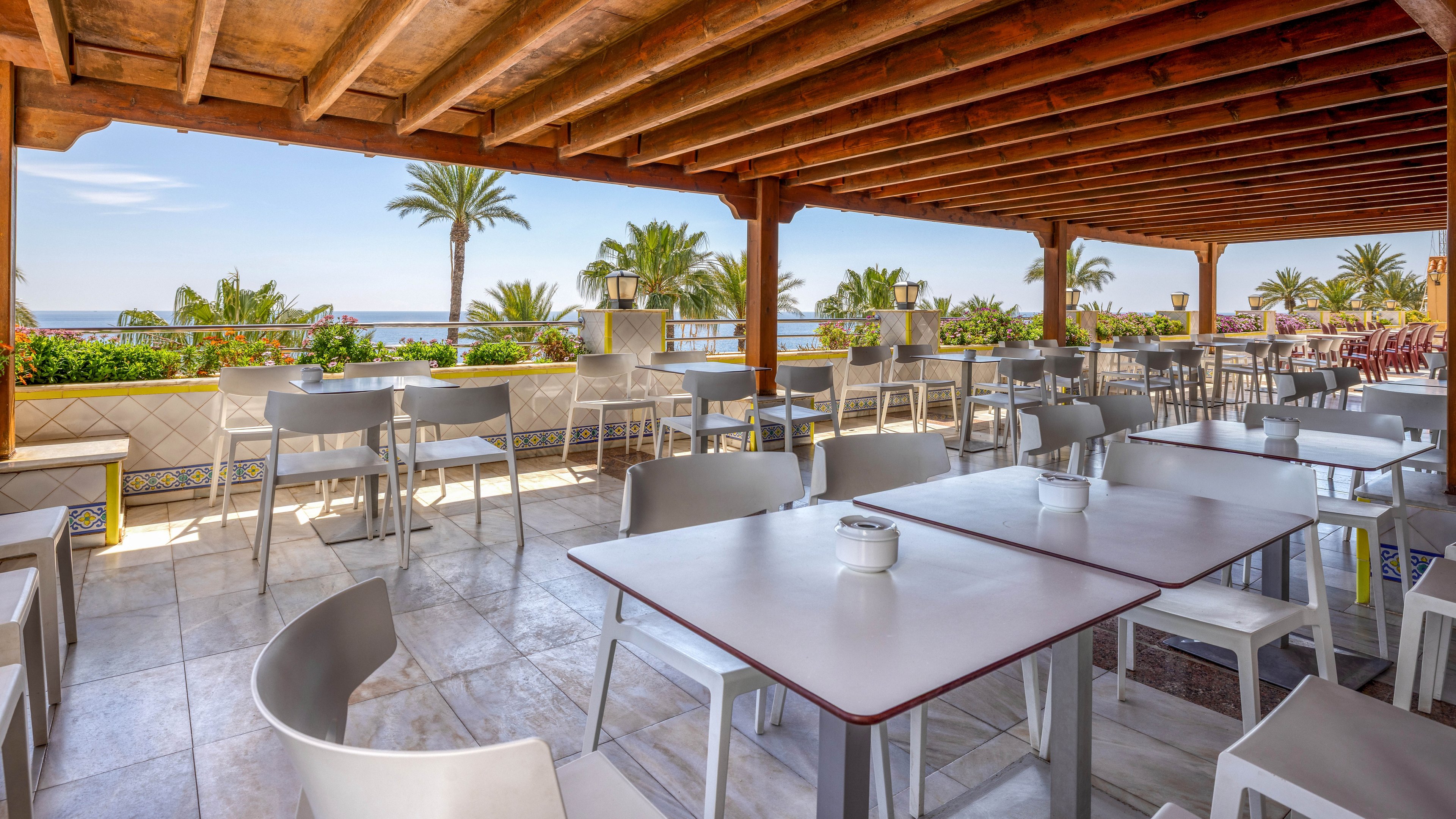 An open-air terrace café features neatly arranged white tables and chairs under a wooden pergola roof. The seating area overlooks a lush row of plants and palm trees with a clear view of the ocean in the background. Bright daylight illuminates the tiled floor and clean, minimalist furniture. The scene suggests a relaxed coastal dining or coffee setting with no people present.
