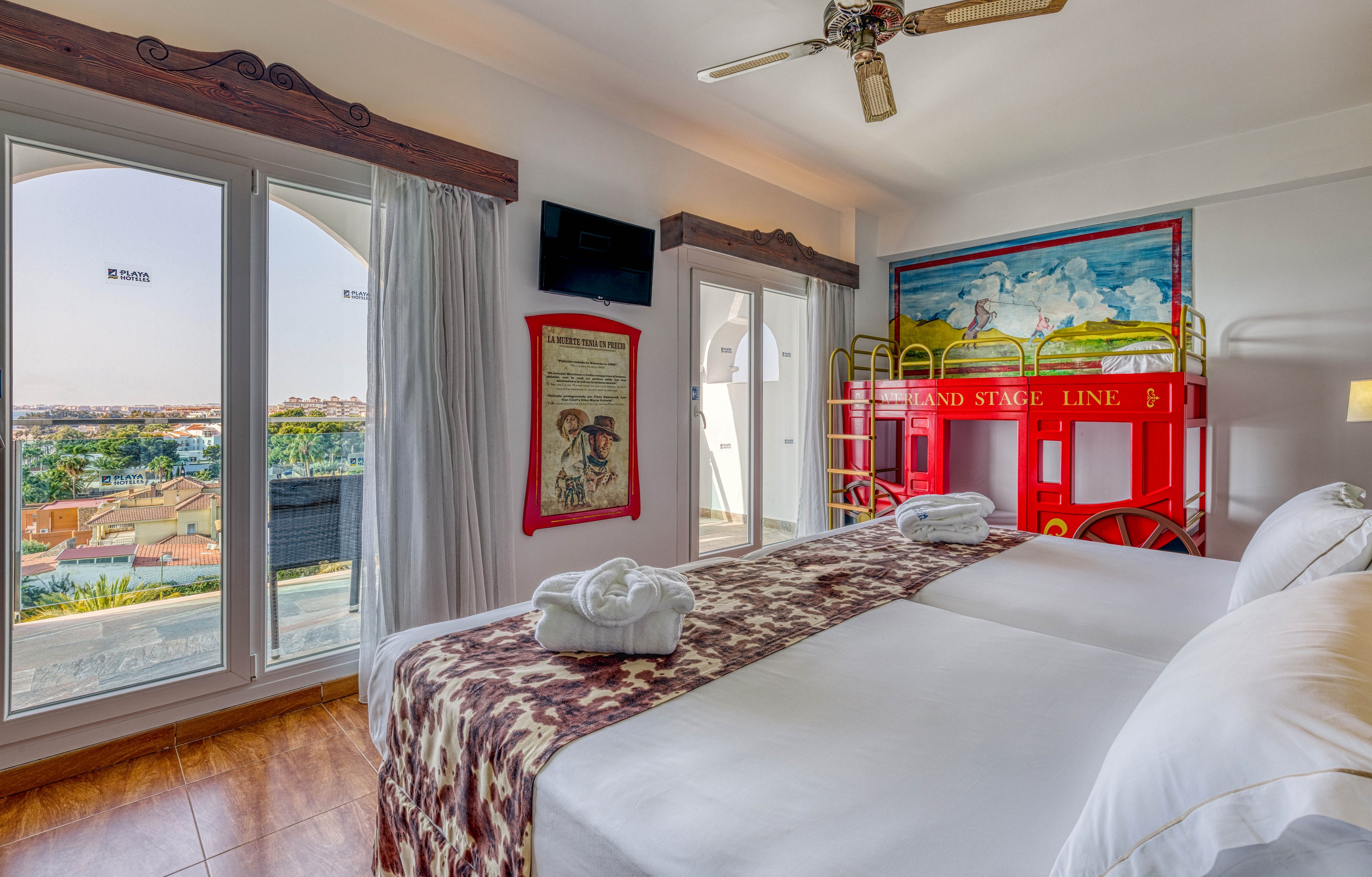 A sunlit hotel room features two neatly made twin beds with patterned throws and folded bathrobes on top. In the corner, a colorful bunk bed designed like a red stagecoach adds a playful, Western-inspired touch. Large windows and a glass door open to a balcony with expansive views of a town and distant landscape. Wall art and a mounted television complete the cozy yet family-friendly interior.
