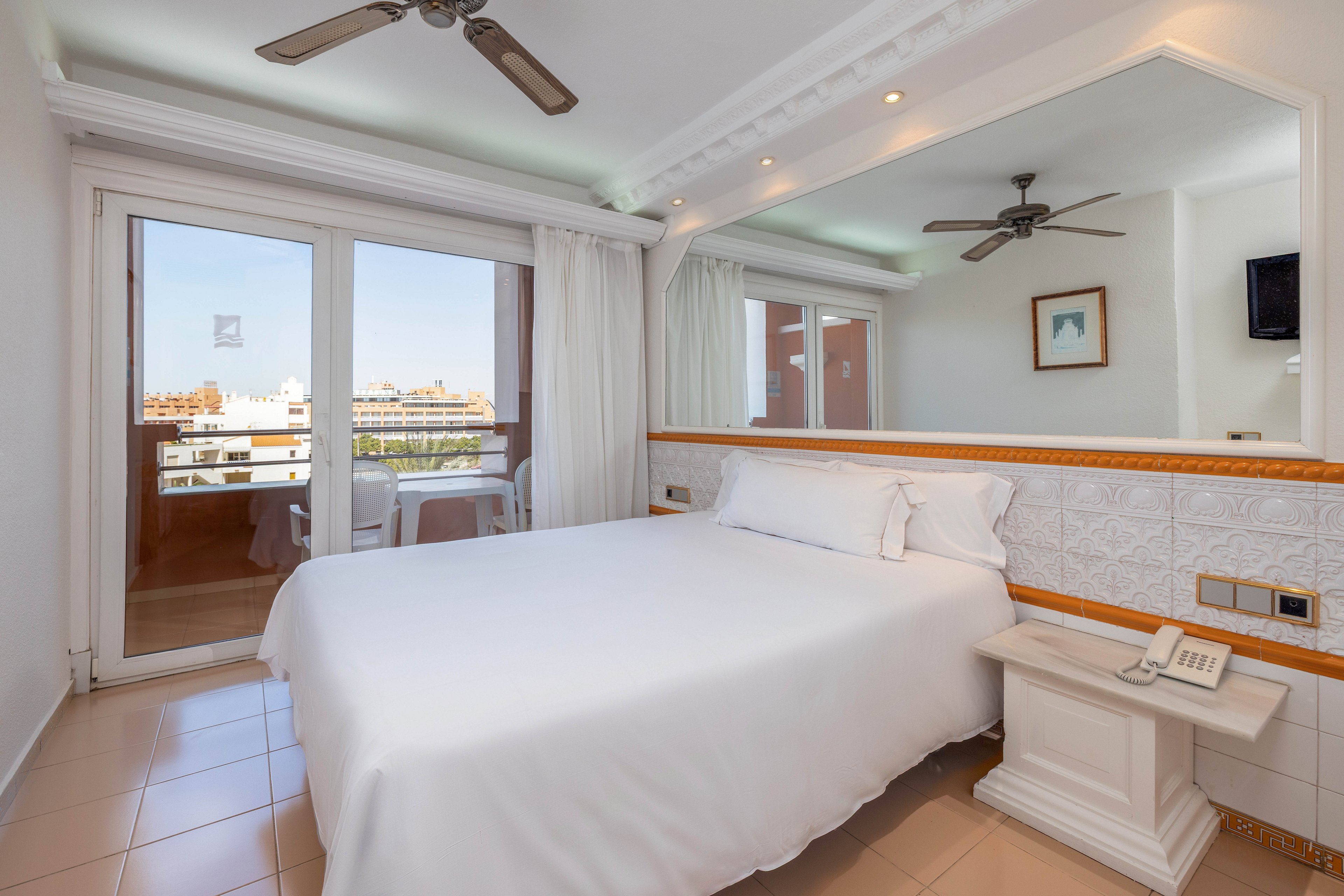 A neatly made double bed with white linens is the focal point of this bright hotel bedroom. Large glass doors open onto a balcony overlooking a cityscape under a clear sky. A wide wall mirror reflects the room, including a ceiling fan and framed artwork. Neutral tiles and soft white curtains contribute to the clean, minimalist aesthetic.