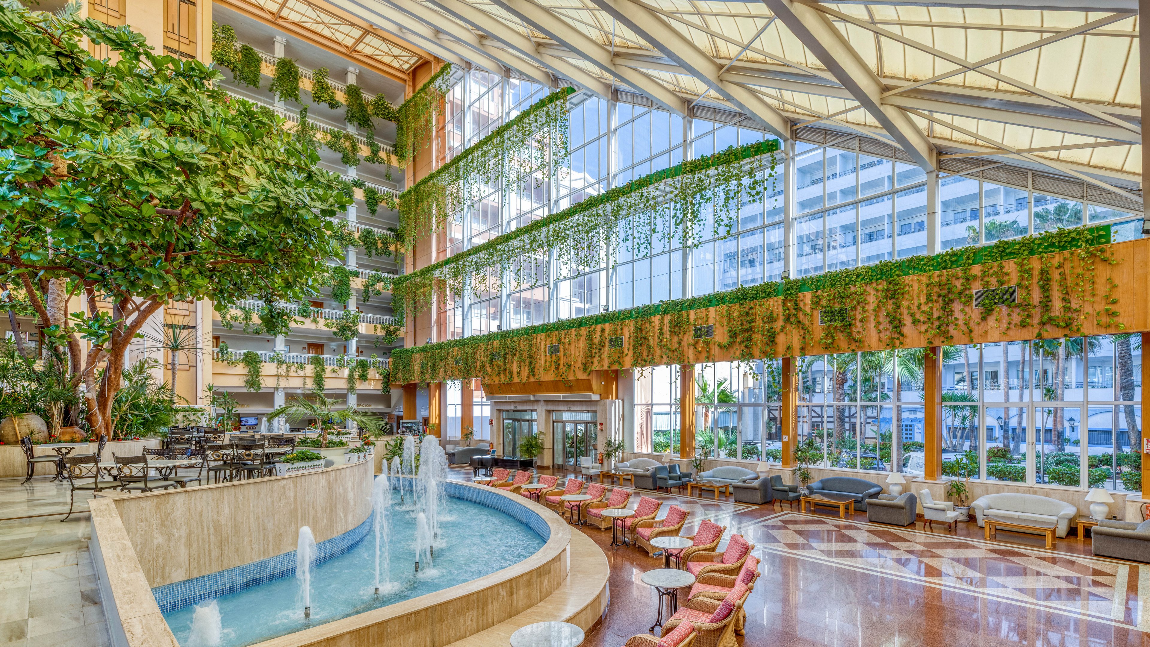 A large, modern hotel atrium features a curved indoor fountain as its central focal point. Comfortable seating areas with armchairs and small tables line the polished floor, creating a relaxed lounge environment. Tall glass windows reveal city buildings outside while allowing abundant natural light to flood the space. Greenery and hanging plants cascade from upper levels, adding a lush, tranquil atmosphere to the bright interior.