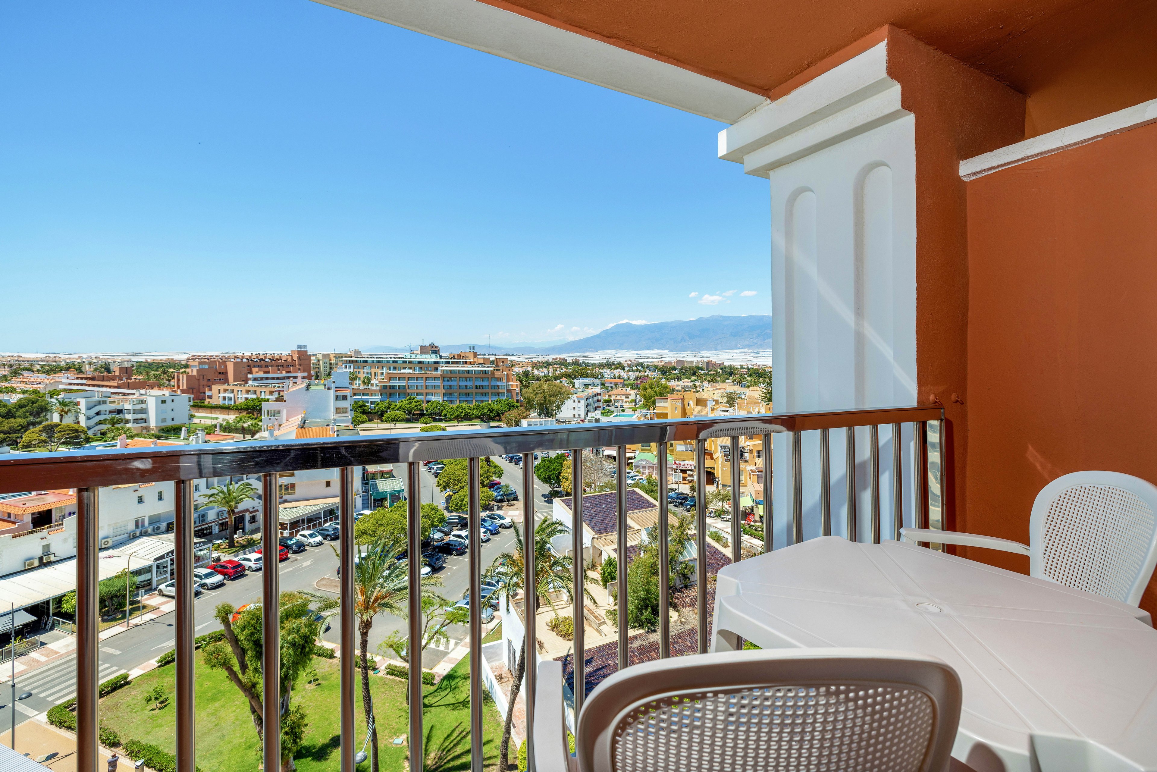 A bright hotel balcony offers a view over a coastal cityscape. White plastic chairs and a table sit on the balcony behind a metal railing. Below, palm trees, streets, and low-rise buildings stretch toward the horizon. In the distance, mountains and a clear blue sky create a relaxed vacation atmosphere.