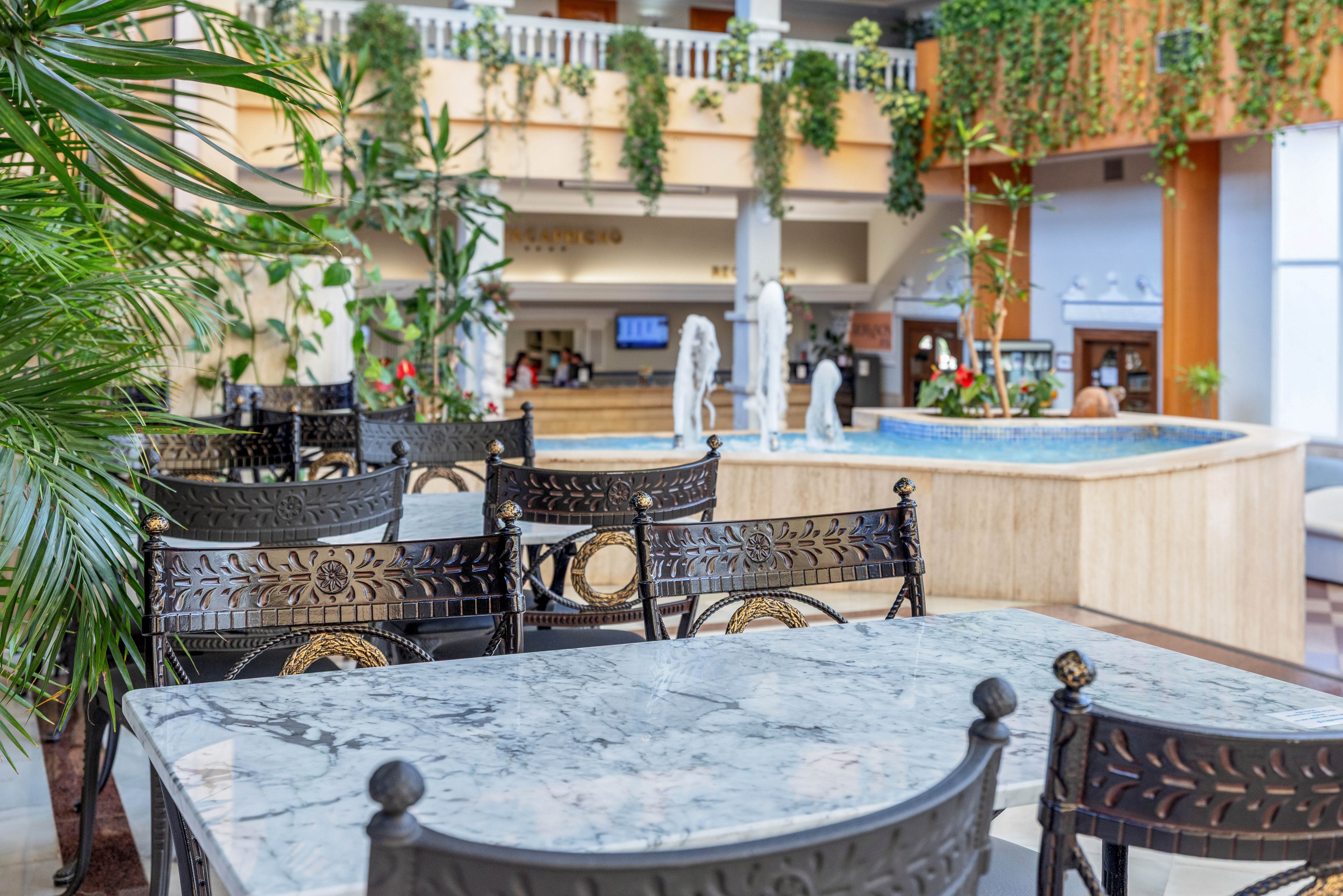 An indoor hotel courtyard features marble-topped tables and ornate metal chairs in the foreground. A decorative fountain with sculpted figures stands in the center of the space. The background shows a reception or bar area with digital screens and shelving. Lush green plants cascade from upper balconies and walls, adding a fresh, tropical atmosphere.