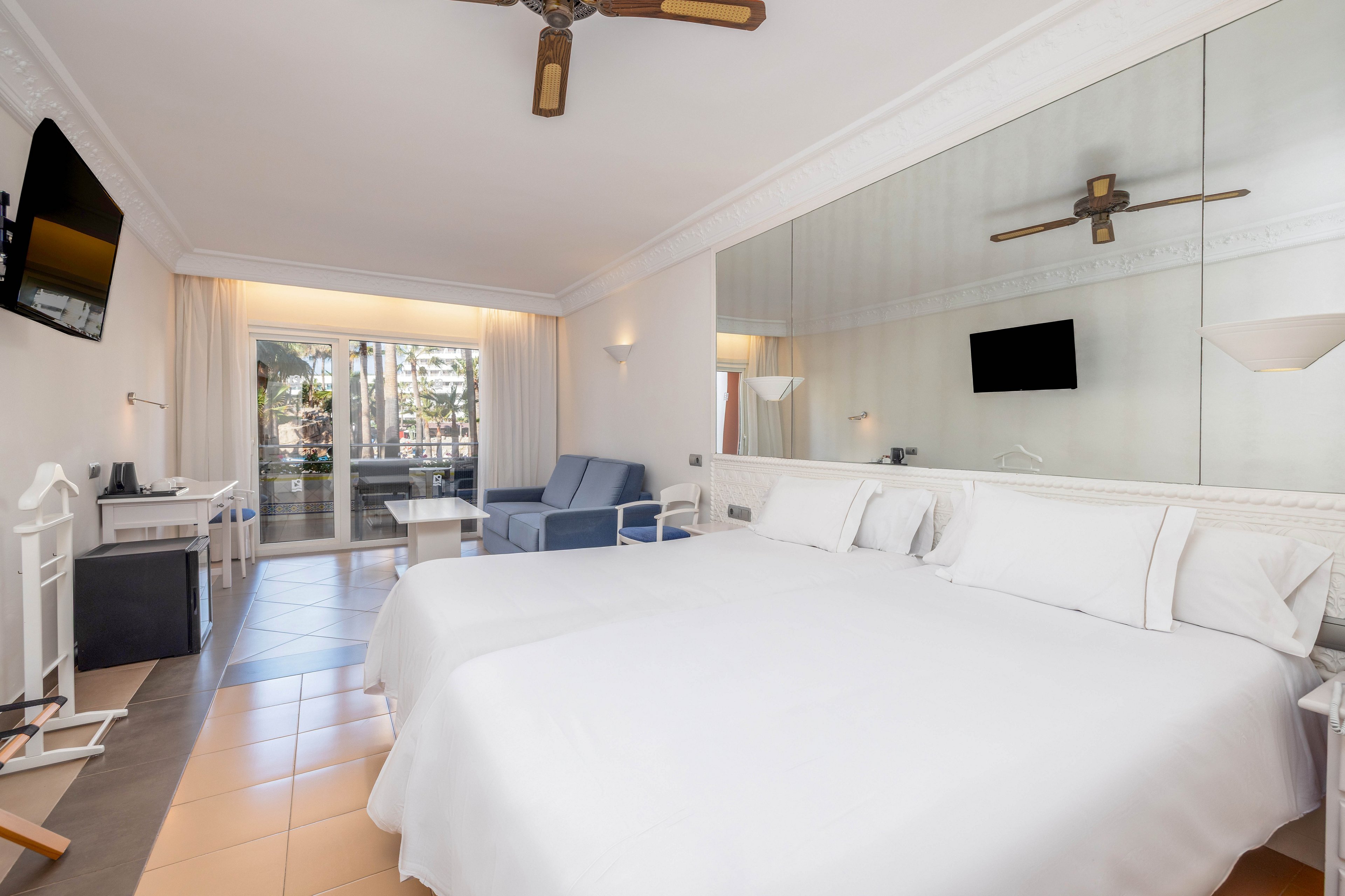 A spacious hotel room features a large neatly made bed with white linens and pillows. The interior includes a wall-mounted television, a desk area, and a blue sofa near the balcony. A large mirror runs along the wall behind the bed, enhancing the sense of space and light. Floor-to-ceiling curtains open to a balcony with outdoor seating and a view of palm trees and a pool area.
