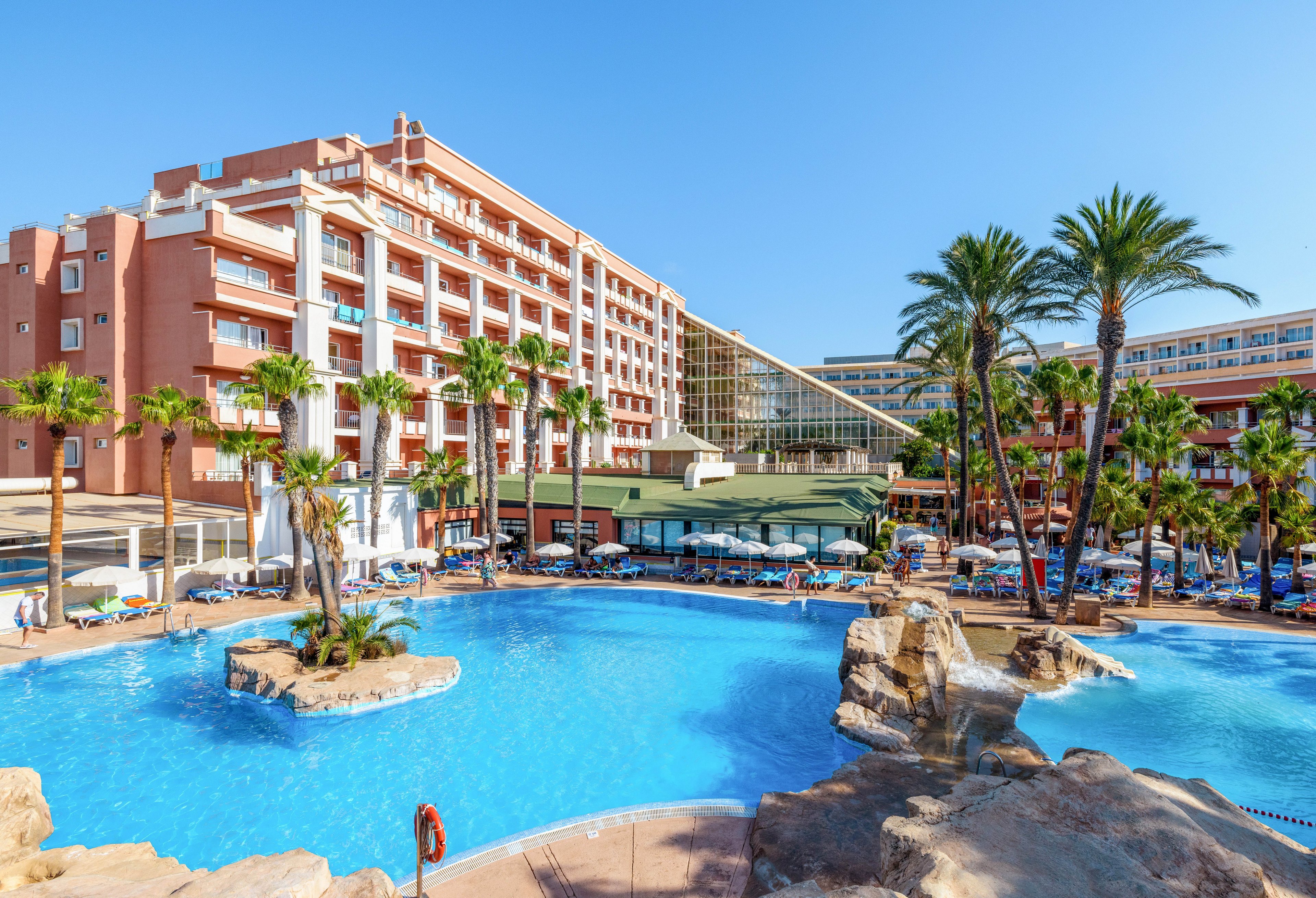 A large resort hotel overlooks a bright blue outdoor swimming pool surrounded by palm trees. Guests relax on sun loungers and under umbrellas around the pool area. The multi-story building features balconies and a modern glass atrium section. Clear blue sky and sunlight create a warm, inviting vacation atmosphere.