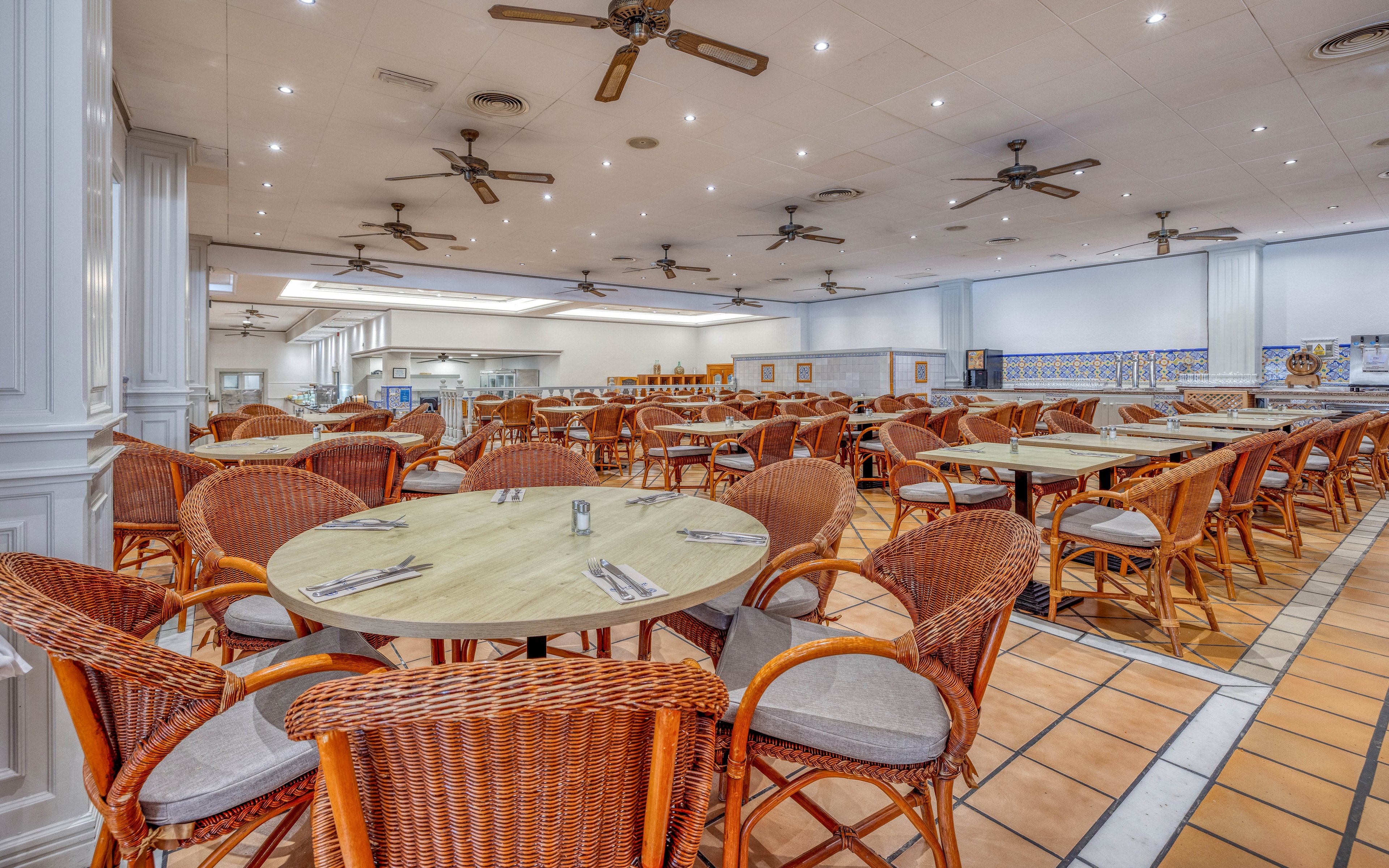 A spacious restaurant dining hall features round wooden tables surrounded by wicker chairs. The room is empty, with neatly arranged cutlery and napkins set on each table. Ceiling fans and recessed lighting line the white ceiling, while tiled floors and white columns add to the clean, airy atmosphere. The open kitchen area is visible in the background, suggesting a buffet or cafeteria-style service.