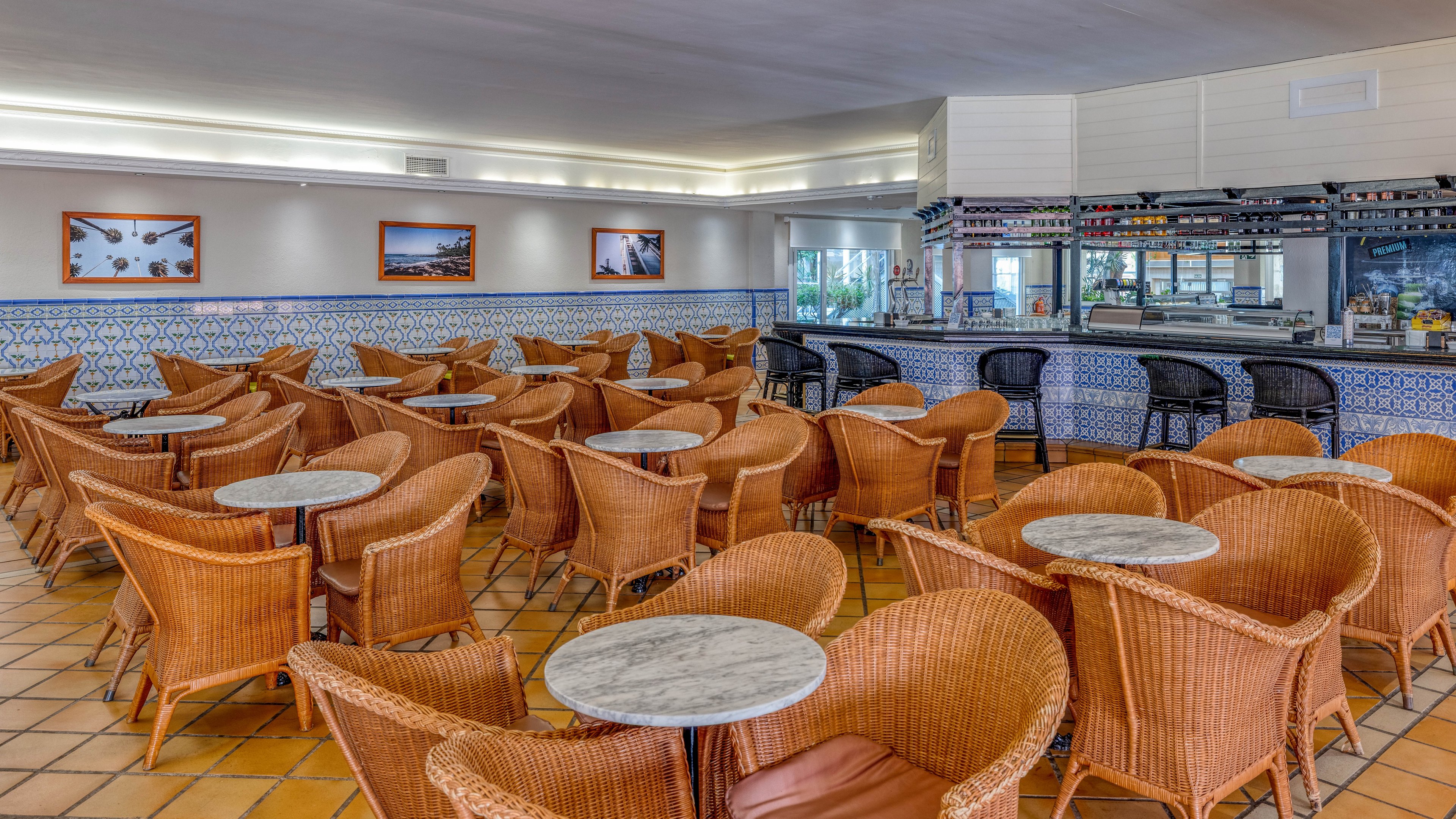 A bright café interior features numerous wicker chairs arranged around small round marble tables. The space is empty of people, highlighting the orderly layout and warm tones of the tiled floor. A long bar with shelves of bottles and glasses runs along one side, accented by patterned blue and white wall tiles. Framed photographs hang on the walls, adding visual interest to the relaxed, coastal-inspired setting.