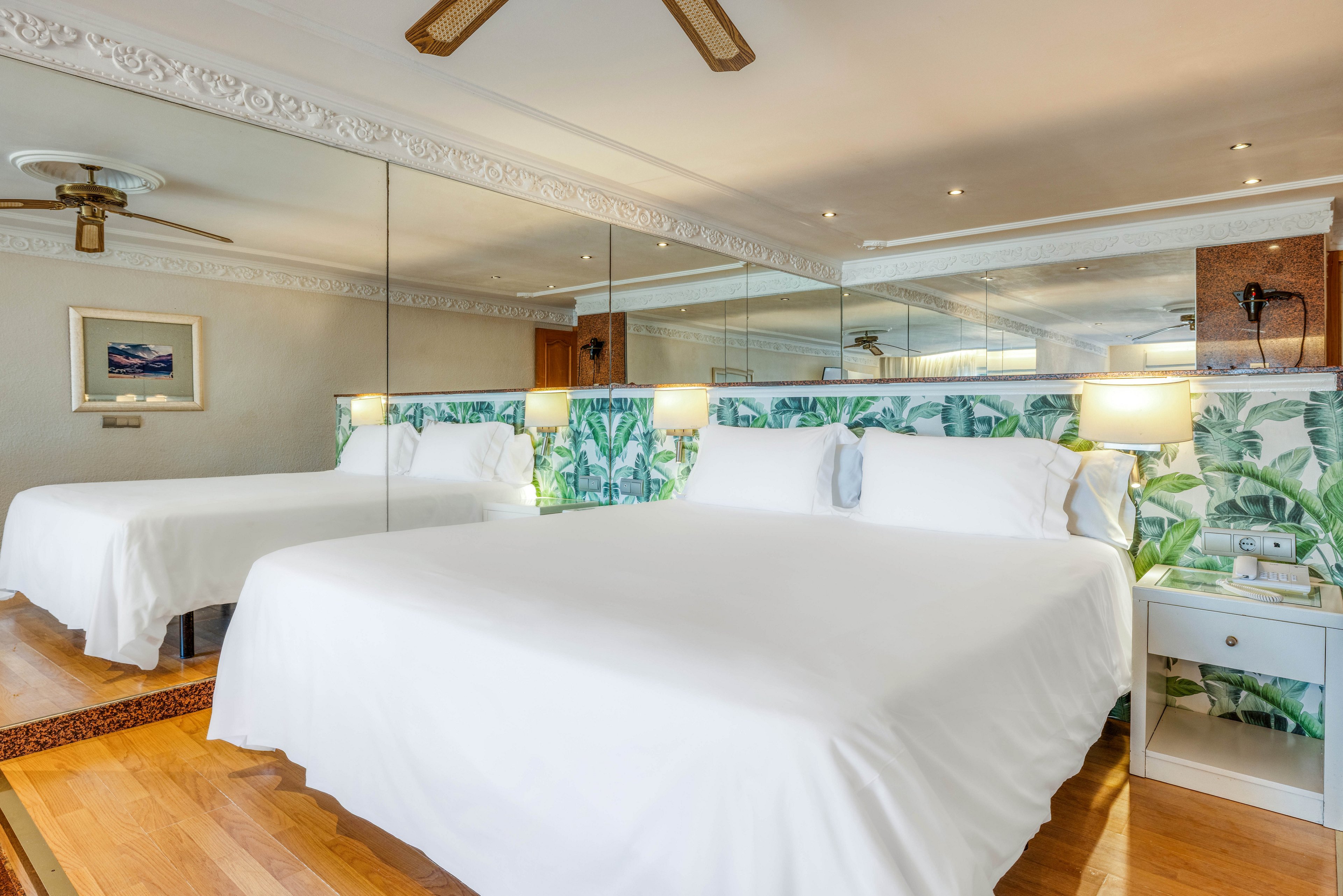 A spacious hotel bedroom features a large white bed centered on a wooden floor. A full mirrored wall behind the bed reflects the room, creating a sense of depth and openness. Tropical leaf-patterned wallpaper runs along the headboard area, adding a fresh, vibrant accent. Soft lighting from bedside lamps and ceiling spotlights creates a warm, inviting atmosphere.