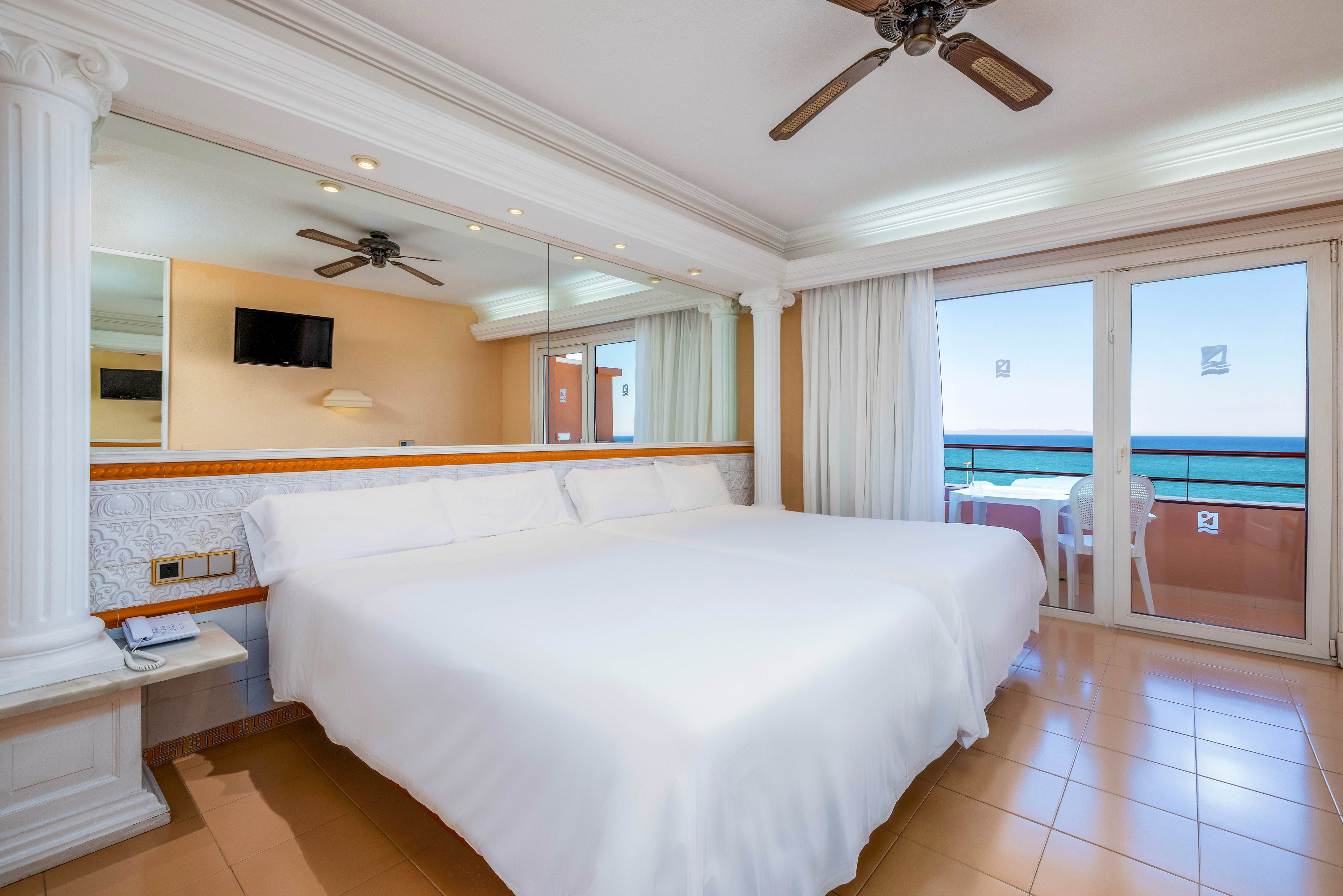 A bright hotel bedroom features a large neatly made bed with crisp white linens. The room includes a wall-mounted television, bedside tables, and a large mirror that reflects the space. Sliding glass doors open onto a balcony with chairs, offering a clear view of the ocean and blue sky. Warm-toned floor tiles and soft lighting create a clean, inviting atmosphere ideal for a beachside stay.