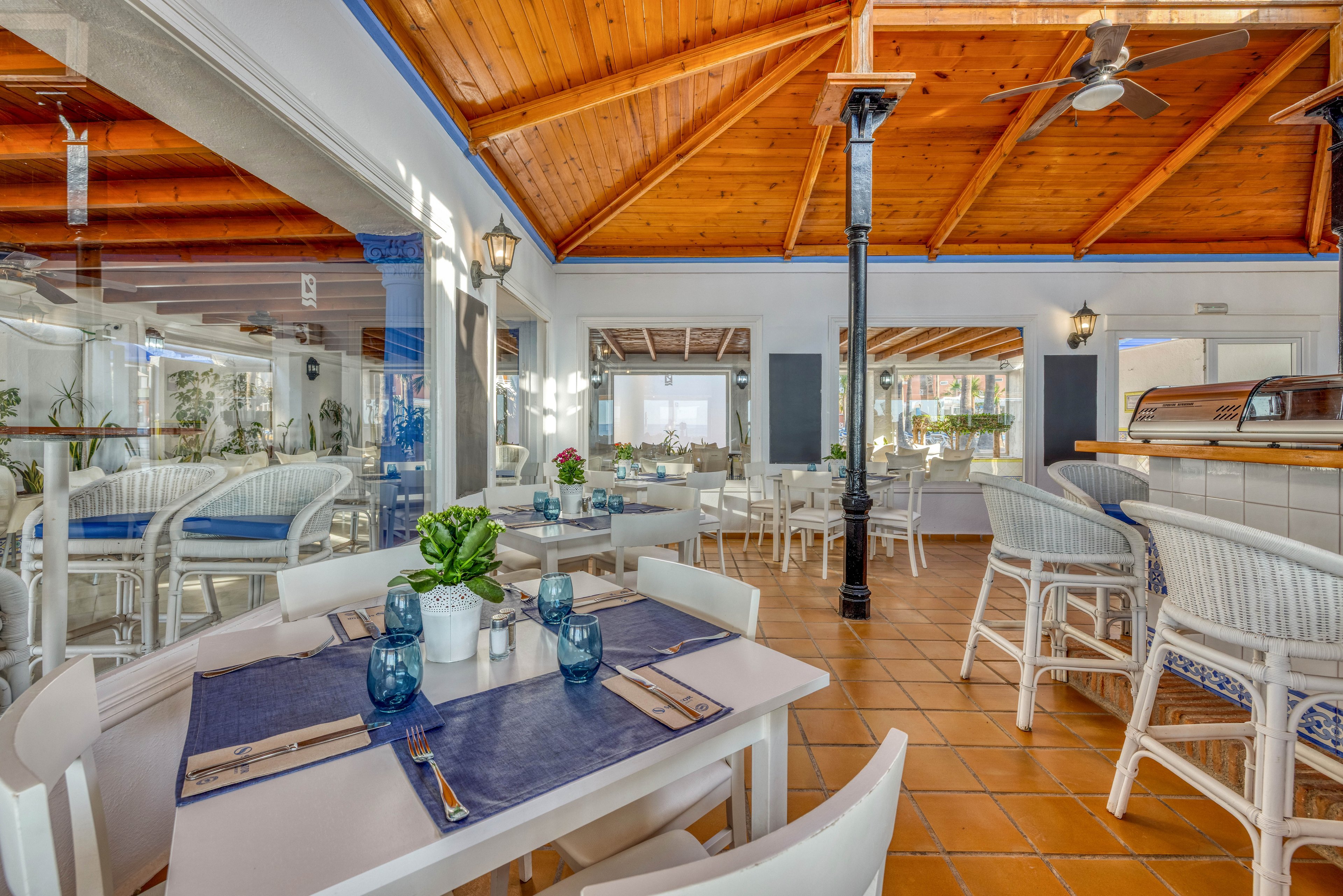 A bright coastal-style restaurant interior features white tables and wicker chairs arranged neatly under a warm wooden ceiling. Blue placemats, glassware, and napkins add a nautical accent to the dining setup. Large windows and mirrors bring in natural light, highlighting the terracotta tile floor and white walls. The space feels airy and inviting, ideal for relaxed seaside dining.