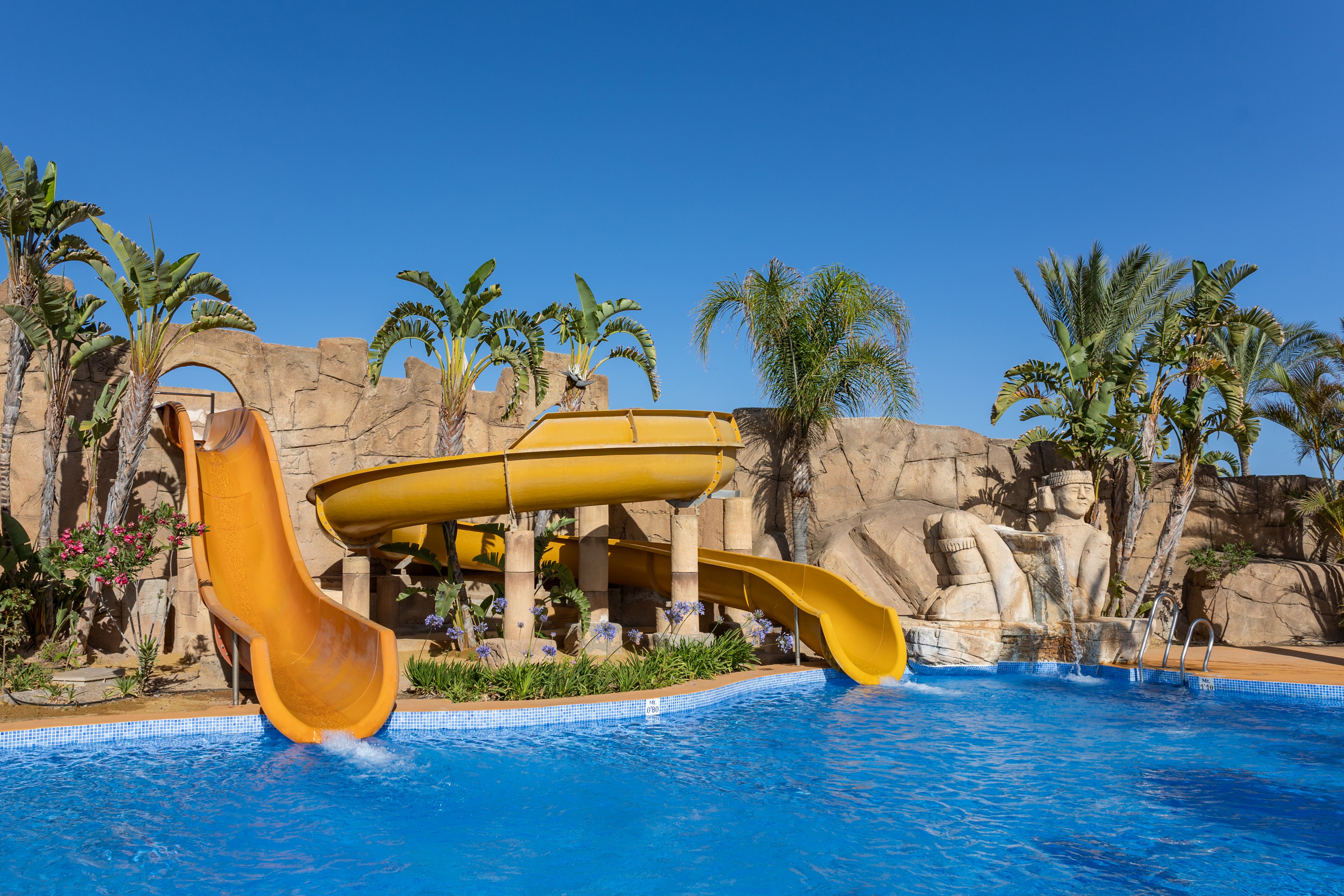 A bright blue swimming pool features large yellow waterslides curving into the water. The slides are set against artificial rock formations with palm trees and lush greenery. Decorative statues and stone structures give the area an exotic, themed resort appearance. The clear sky and vivid colors create a sunny, fun vacation atmosphere.