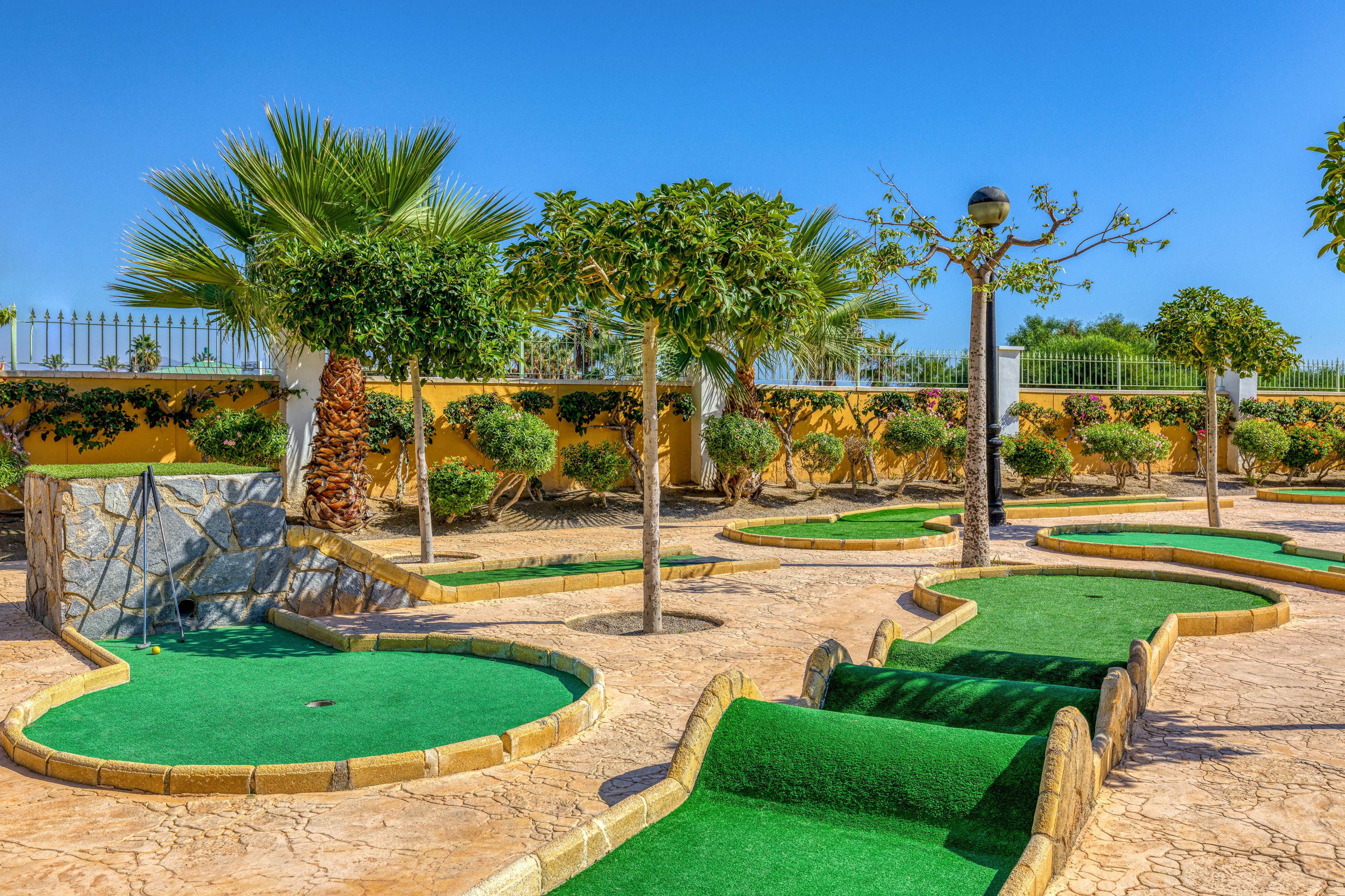 An outdoor mini golf course is shown under a bright blue sky. Curved putting greens with artificial turf wind through a sunlit, stone-textured surface. Palm trees and trimmed shrubs line the course, adding a tropical feel. A low fence and distant greenery frame the background, suggesting a leisure or resort setting.
