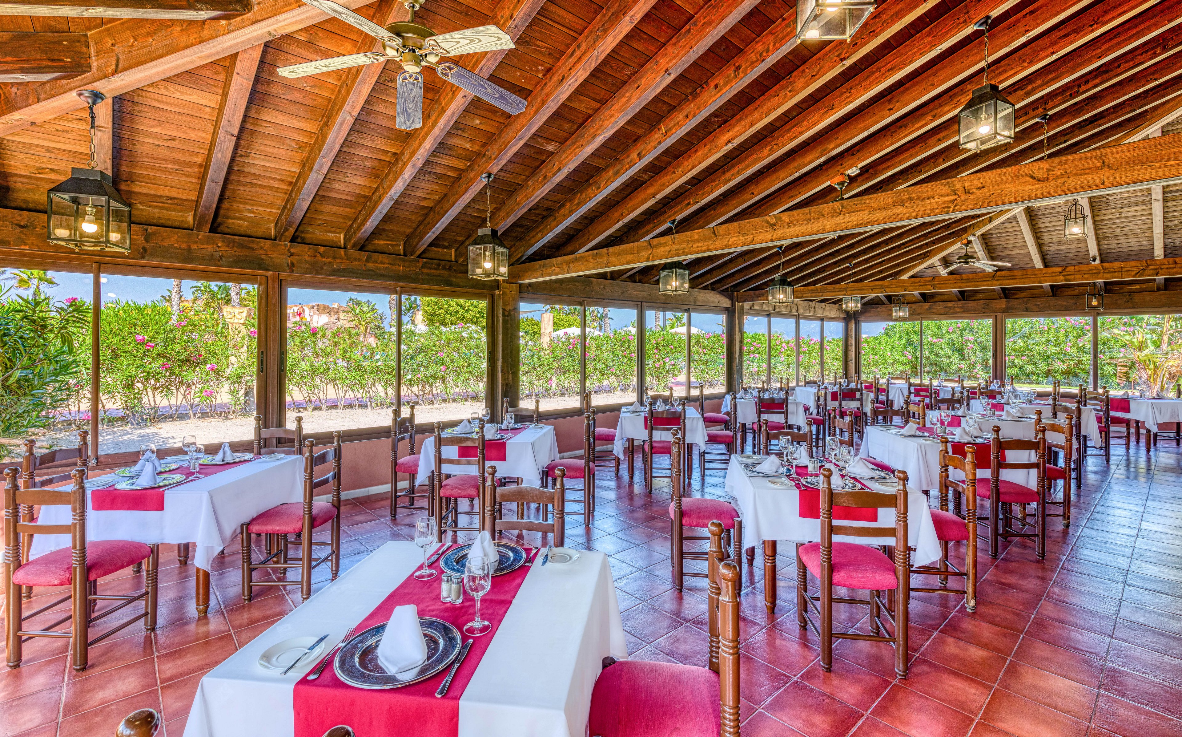 Spacious open-air restaurant dining area with neatly arranged tables and chairs. White tablecloths with red runners and red seat cushions create a vibrant, coordinated look. The wooden ceiling with exposed beams, ceiling fans, and hanging lanterns adds rustic charm. Large open windows reveal lush greenery outside, suggesting a relaxed, resort-style setting.