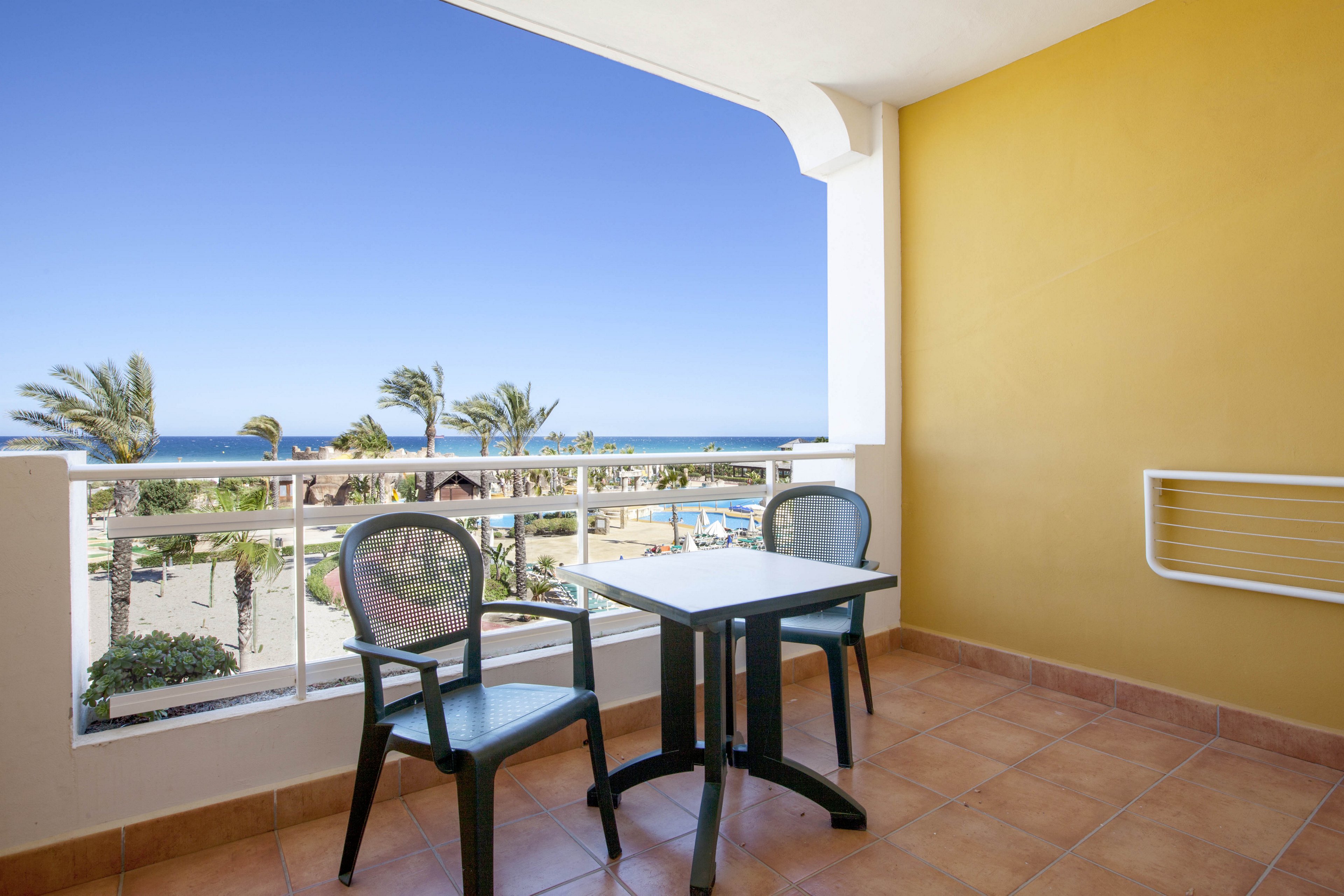 A bright hotel balcony features a small square table and two plastic chairs overlooking a coastal resort. Beyond the white railing, palm trees and a swimming pool area lead toward the blue ocean and clear sky. The tiled floor and yellow wall create a warm, Mediterranean feel. The scene suggests a relaxing vacation spot with outdoor seating and sea views.