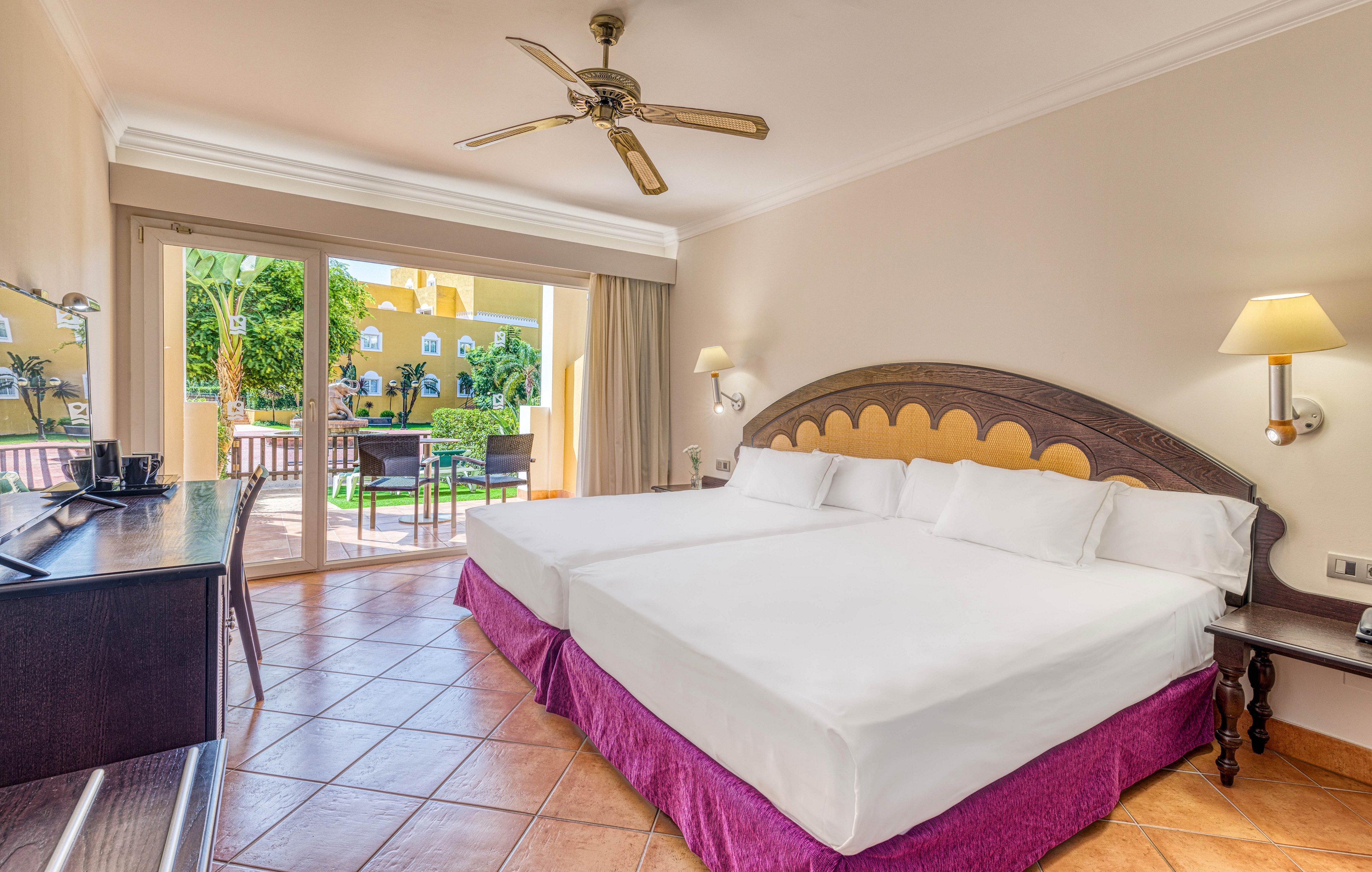 Spacious hotel room featuring two large beds with white linens and purple bed skirts. The room opens onto a terrace with outdoor chairs and a view of a sunny courtyard and palm trees. Warm tile flooring, a ceiling fan, and soft wall lighting create a comfortable vacation atmosphere. A desk area with a television and coffee station completes the modern guestroom setup.